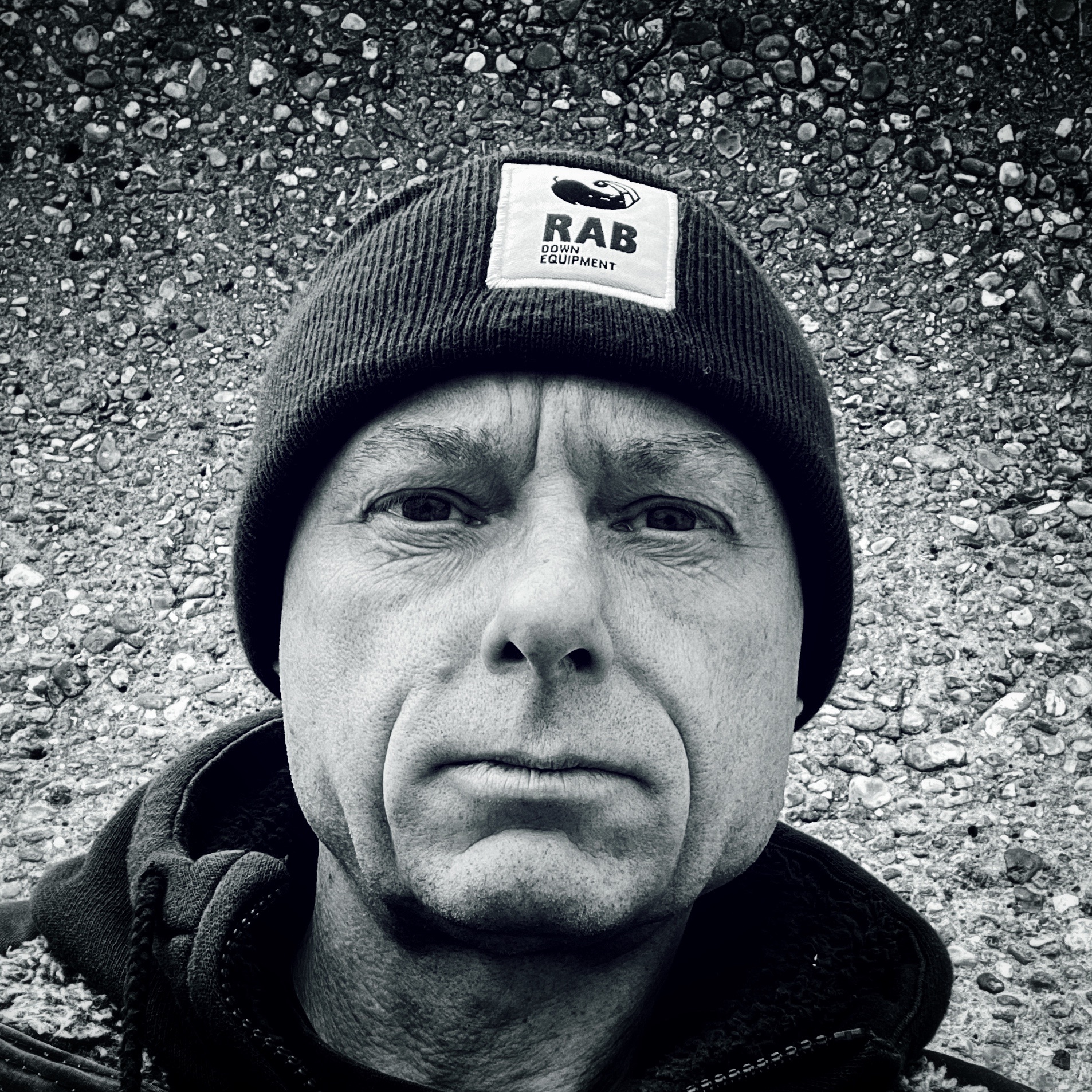 Photo of David Hawkins-Weeks in black and white, wearing a beanie hat, set against a concrete wall