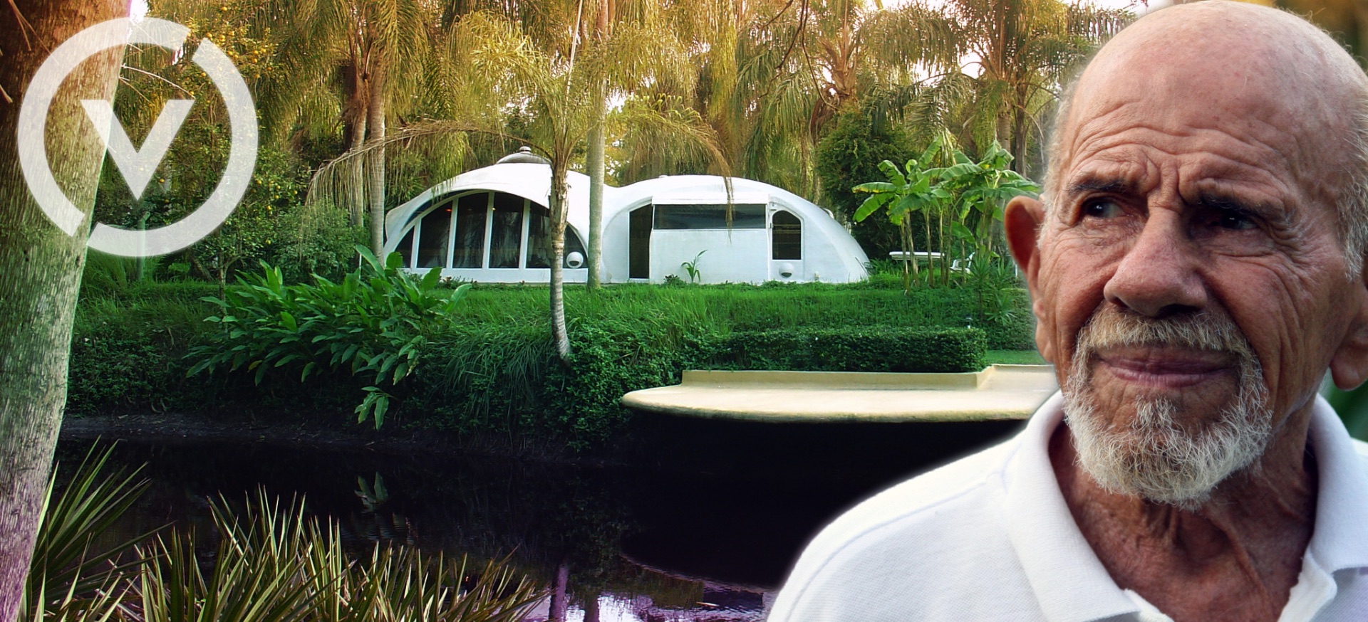 Jacque Fresco beside the Venus Project grounds, with its emblem and buildings among trees.