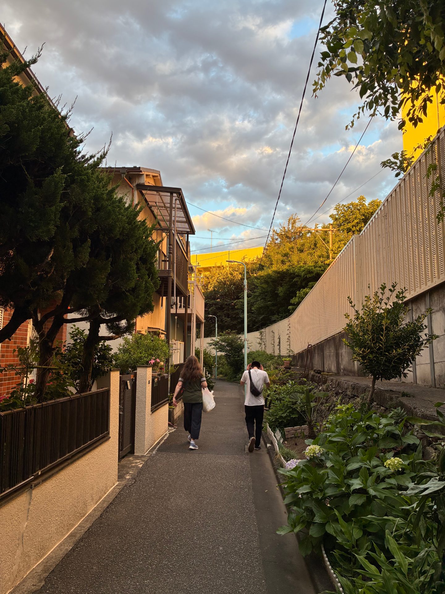 My wife and our son walking down a small back alley in Tokyo during golden hour. The sun is bathing trees and buildings in a soft golden light. She's carrying a bag. There are homes to the left and plants to the right of the foot path. The sky is cloudy with patches of blue peeking through.