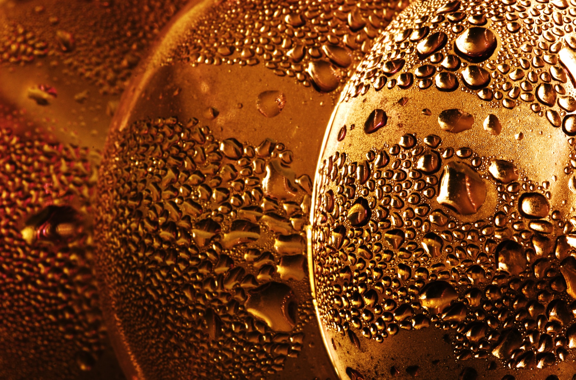 A close-up photograph displays three overlapping, rounded plastic surfaces shimmering with a rich golden-bronze hue. Countless water droplets of various sizes cover the surfaces, catching the light and creating a textured pattern of highlights and shadows. The shallow depth of field keeps the rightmost sphere in sharp focus, while the surfaces behind it gradually blur into the background.