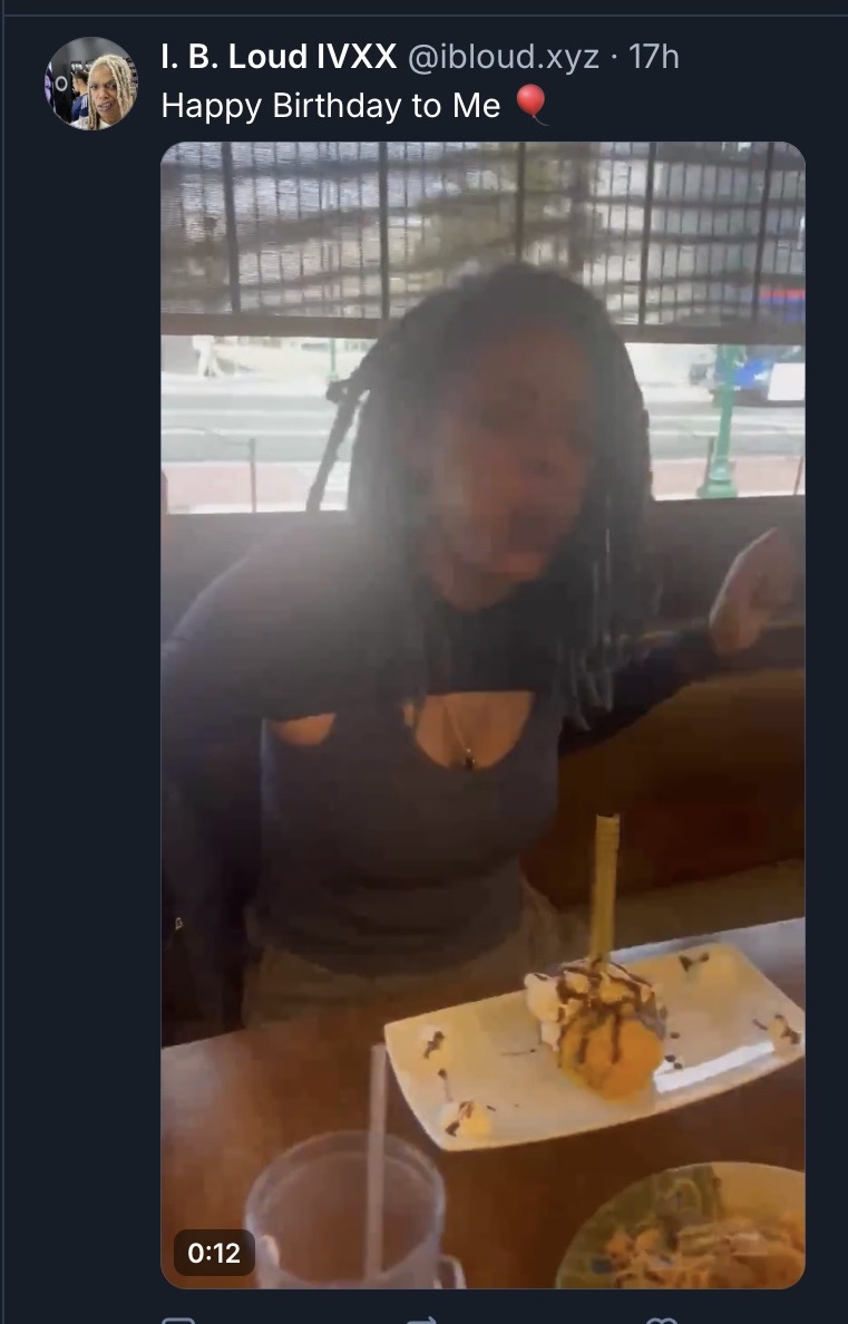 A Black woman with long locs sits at a restaurant table, smiling in anticipation. In front of her is a dessert on a white plate with a single unlit sparkler candle. A drink and a plate of food are also on the table. A window with a street view is visible behind her.
