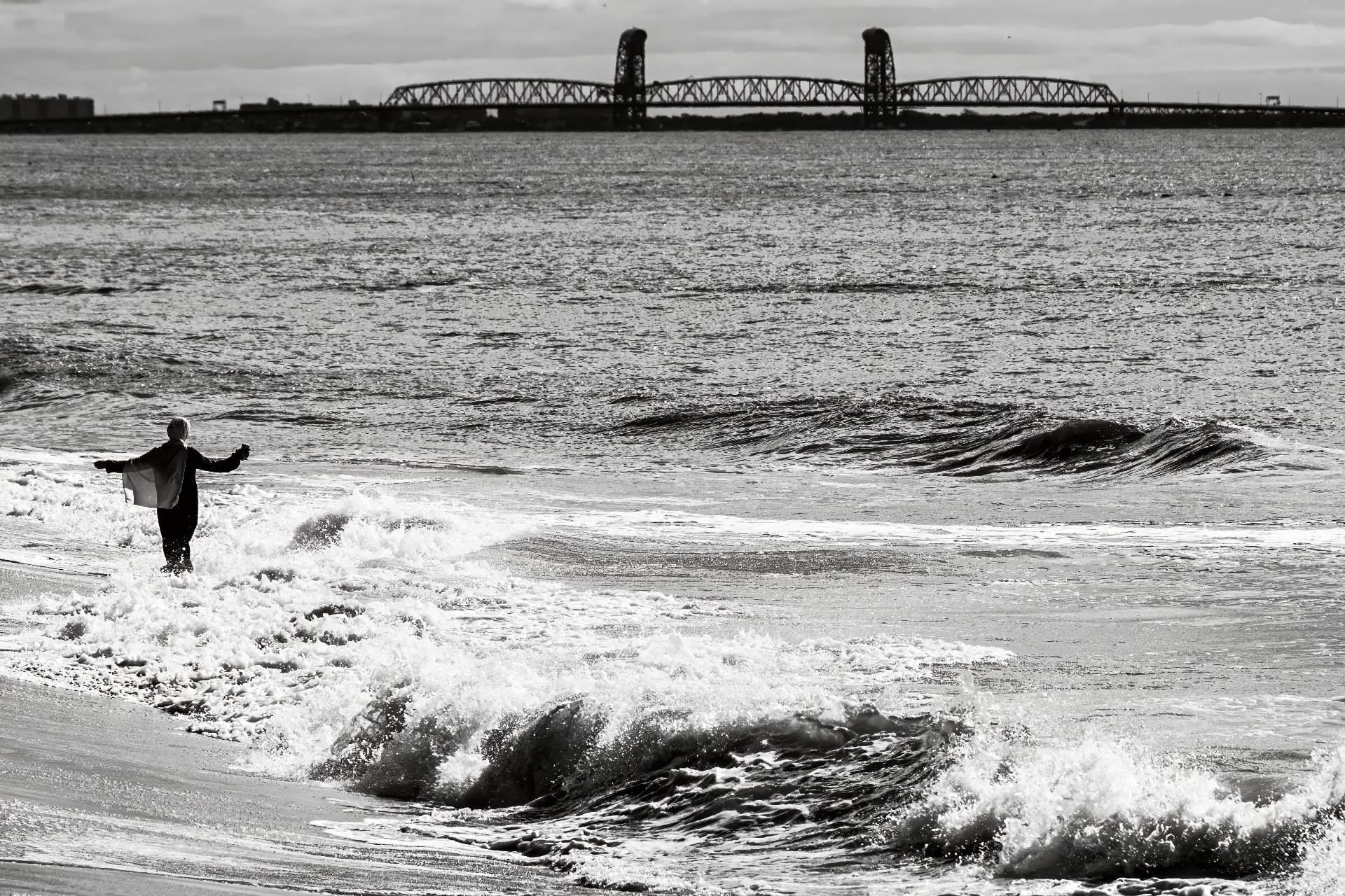 Depicted in black and white, a young woman wearing an abaya and hijab wades through the surf with her arms outstretched at the edge of the beach at Coney Island. The Marine Parkway Bridge can be seen in the background off in the distance.