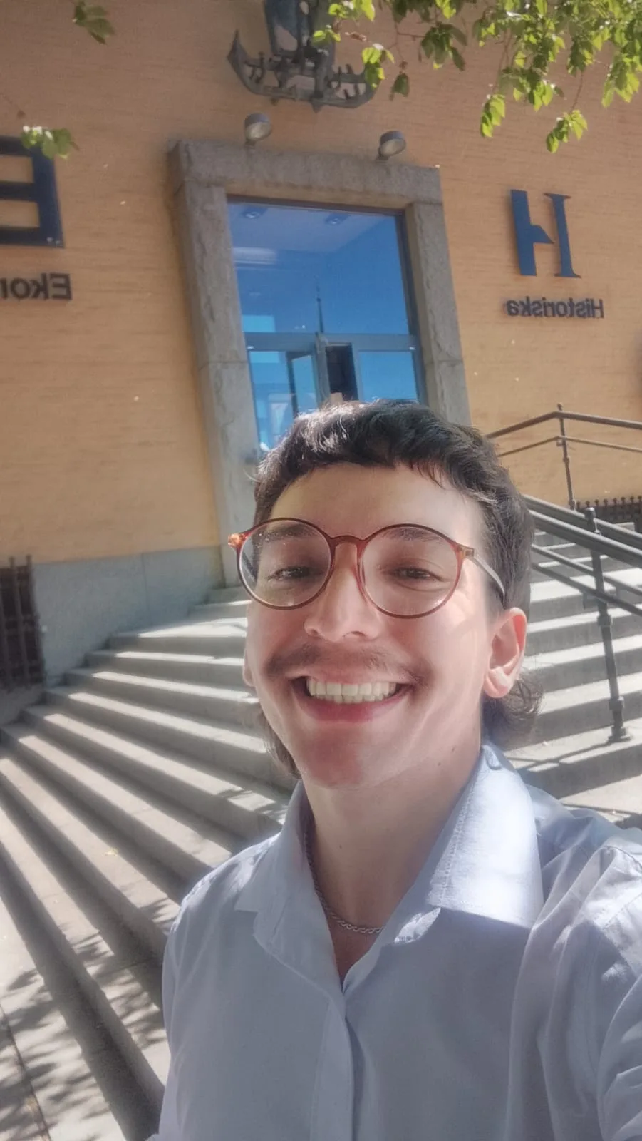 Selfie de Vic Martins, uma pessoa jovem, não binária, branca de pele clara, sorrindo bastante, com cabelo curto e escuro em corte mullet e óculos de armação redonda castanho-escura. Veste camisa social branca. Ao fundo, a fachada do Historiska Museet, em Estocolmo, com uma escadaria e as palavras "Historiska" visíveis (parcialmente refletidas/espelhadas na foto), em dia ensolarado com galhos de árvore no topo da imagem.