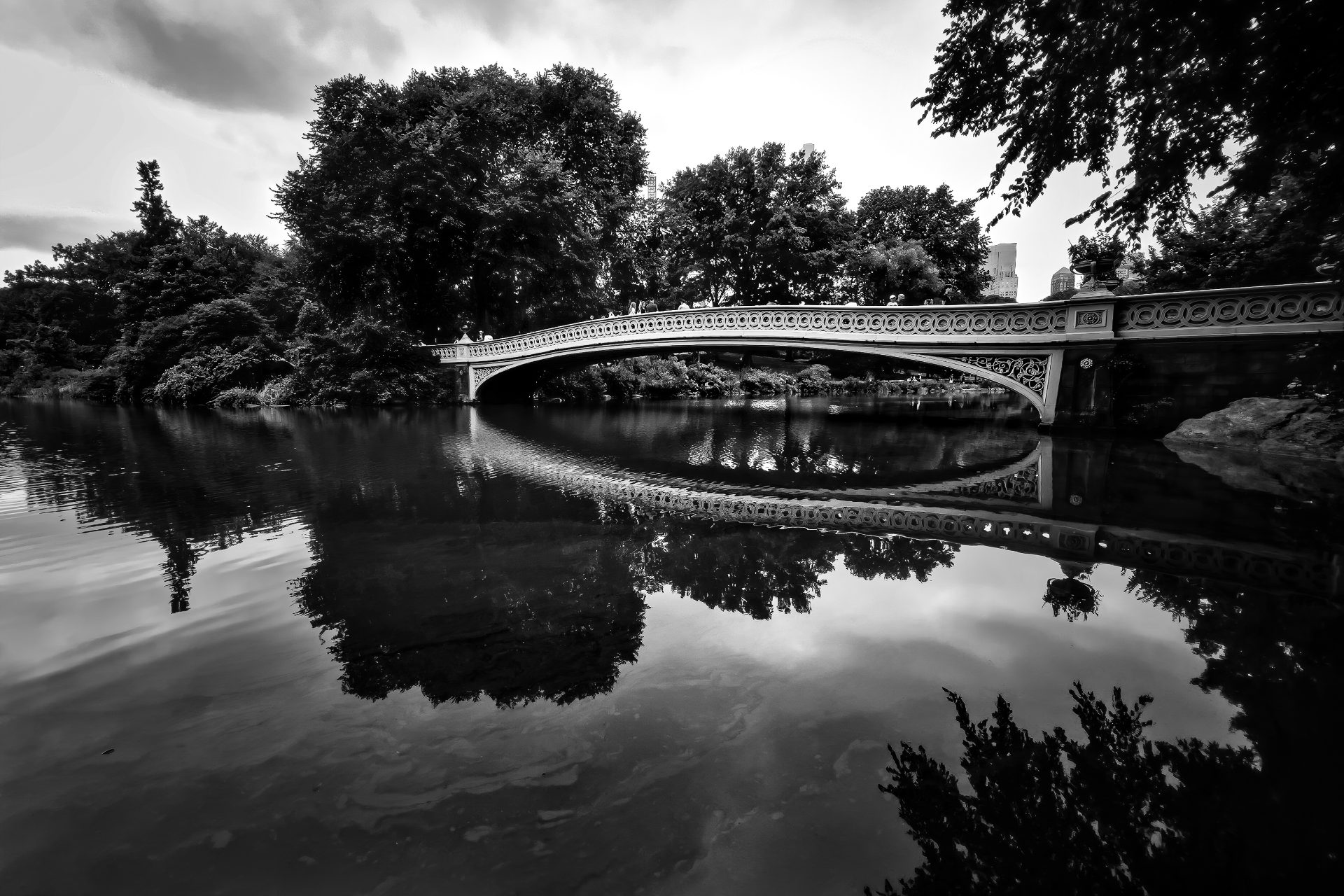 In black and white, the ornate arch of the Bow Bridge and the nearby trees are reflected in the water. The bridge spans a narrow part of The Lake in Central Park, Manhattan.