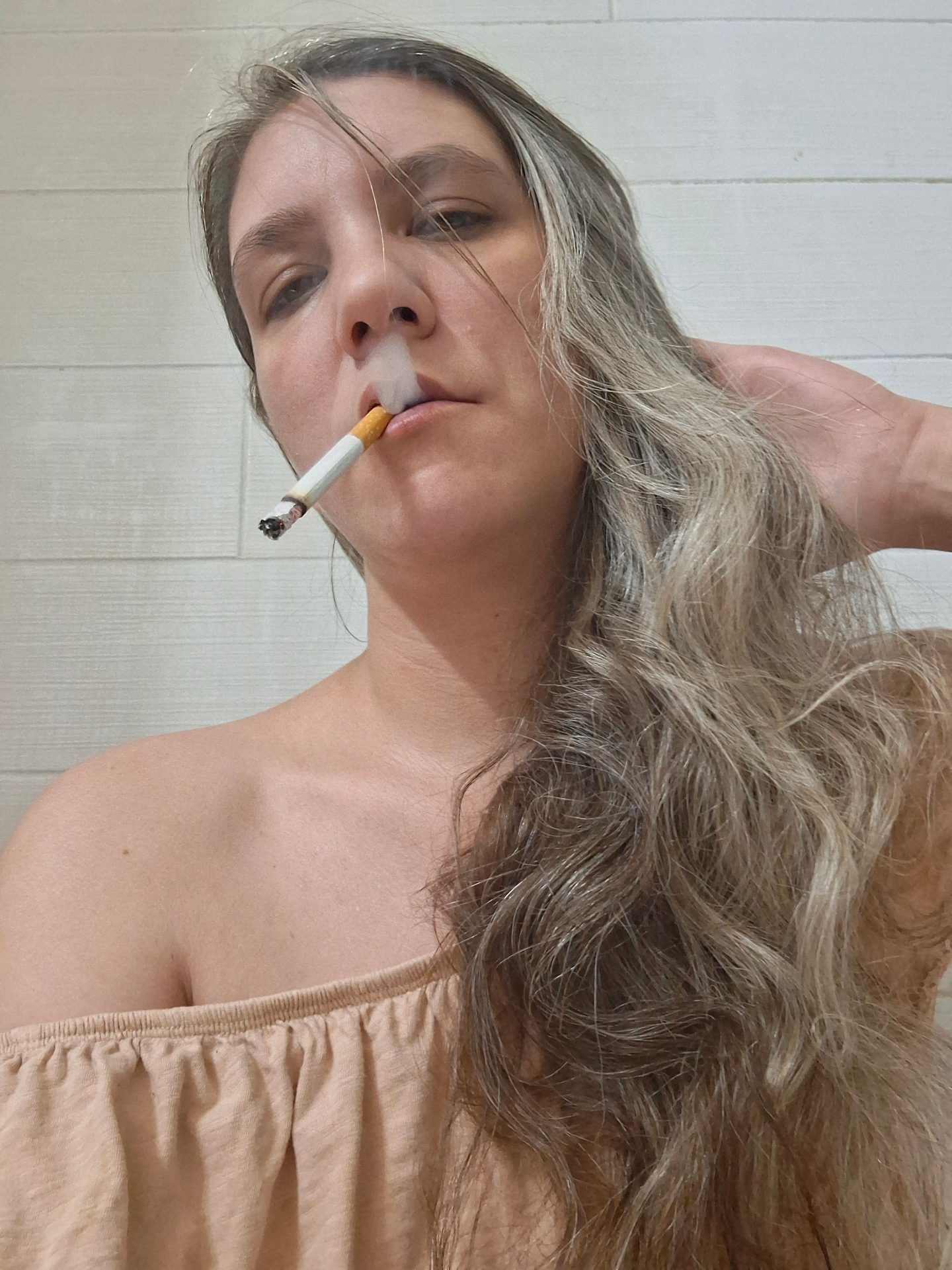 Valerie Rayne begins exhaling a cork-filtered cigarette with her hand in her curly hair.