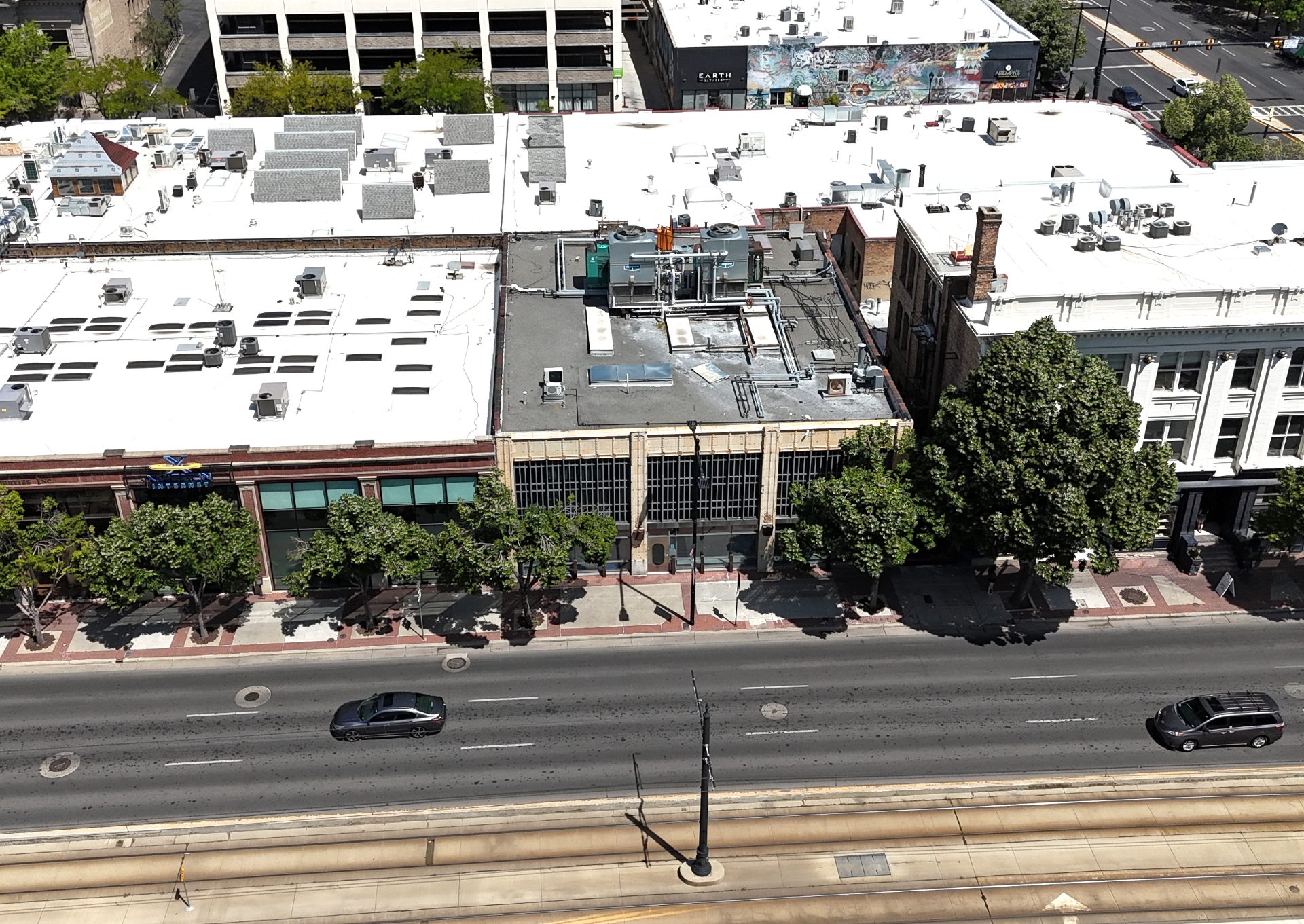 An aerial shot of a street in downtown Salt Lake City. In the center of the frame is a building with tan front and black windows, with large cooling units on the roof. It's surrounded by other low (2-3) story buildings, including the one - directly to the left - that houses XMission. Trees line the street, and the TRAX light rail line can be seen in the foreground.
