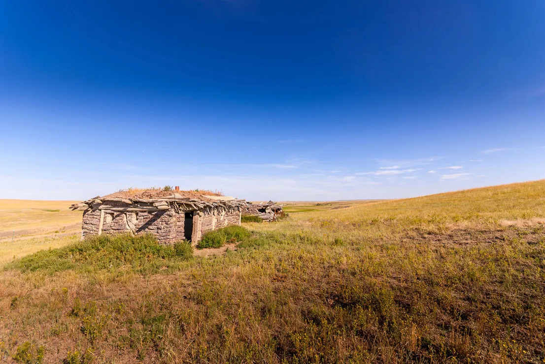A couple of low and dilapidated stone buildings with thatched roofs are the only objects visible under an almost cloudless blue sky in a vast open landscape of rolling hills.