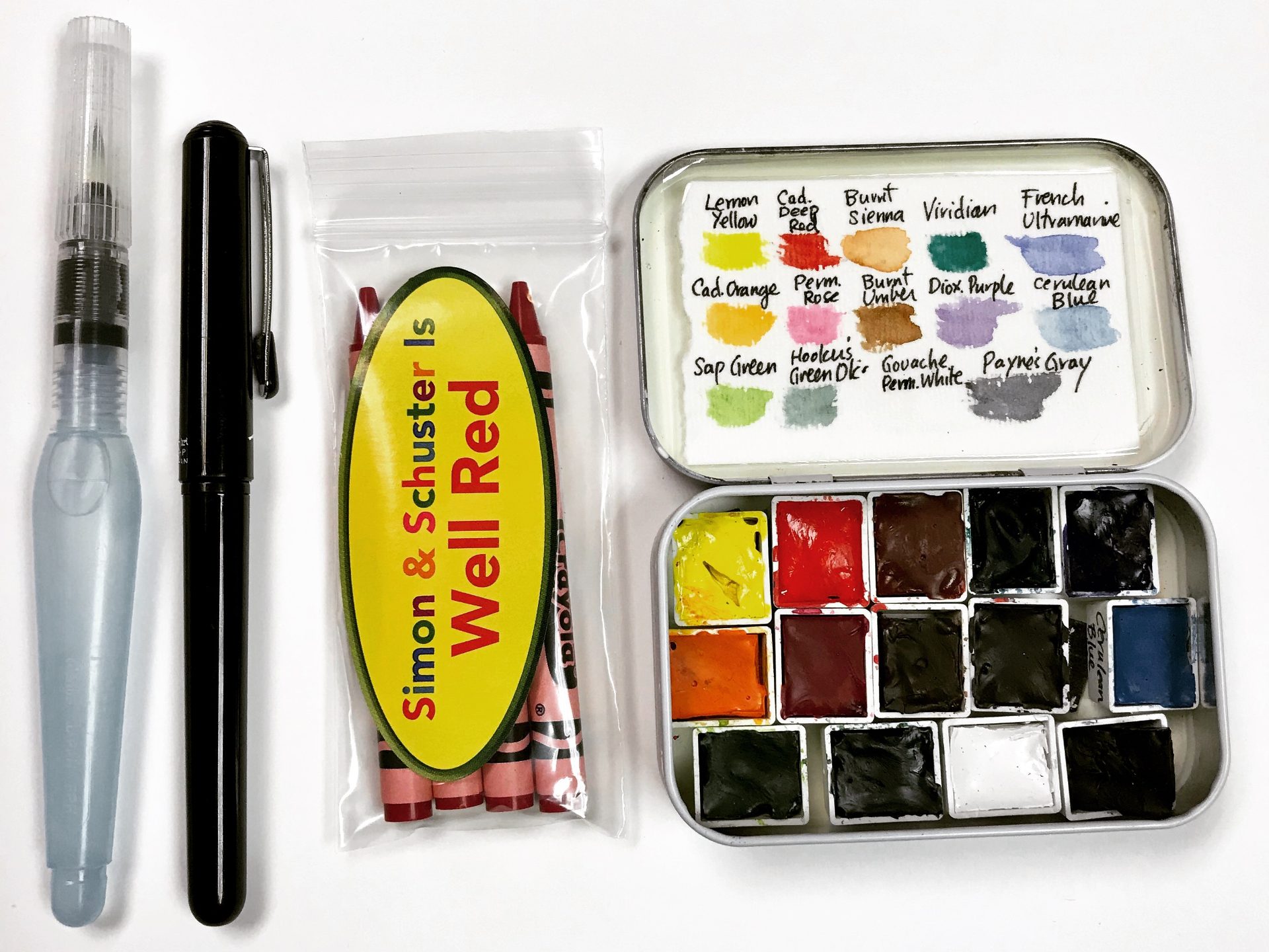 Overhead shot on a white surface featuring a Pentel water brush, a Pentel pocket brush pen, a small plastic bag containing a custom-labeled "Simon & Schuster Is Well Red" crayon package, and a small metal watercolor tin containing a palette of pans with handwritten color labels.