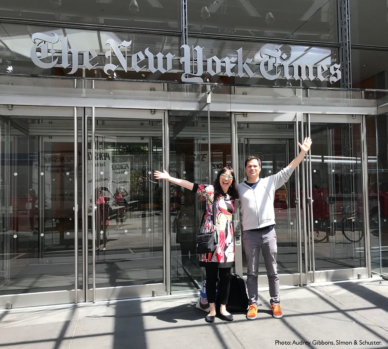 Debbie Ridpath Ohi and Michael Ian Black standing outside the New York Times building with their arms outstretched in front of glass entry doors featuring the New York Times logo. Photo credit: Audrey Gibbons, Simon & Schuster.