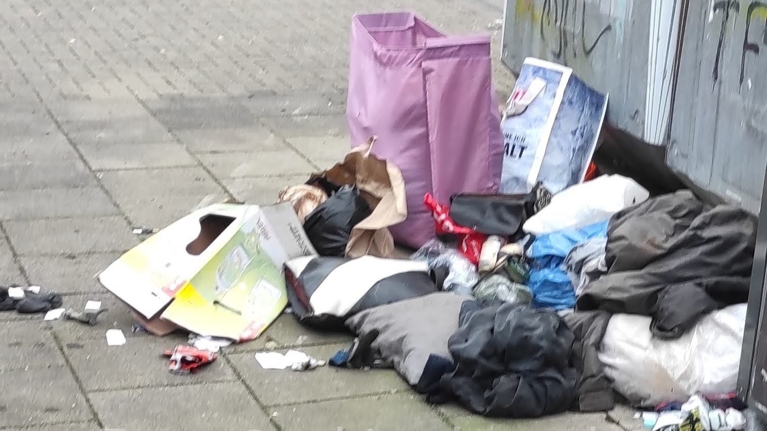 Verschiedene Taschen und aufgerissene Säcke mit Altkleider vor einem Altglascontainer.