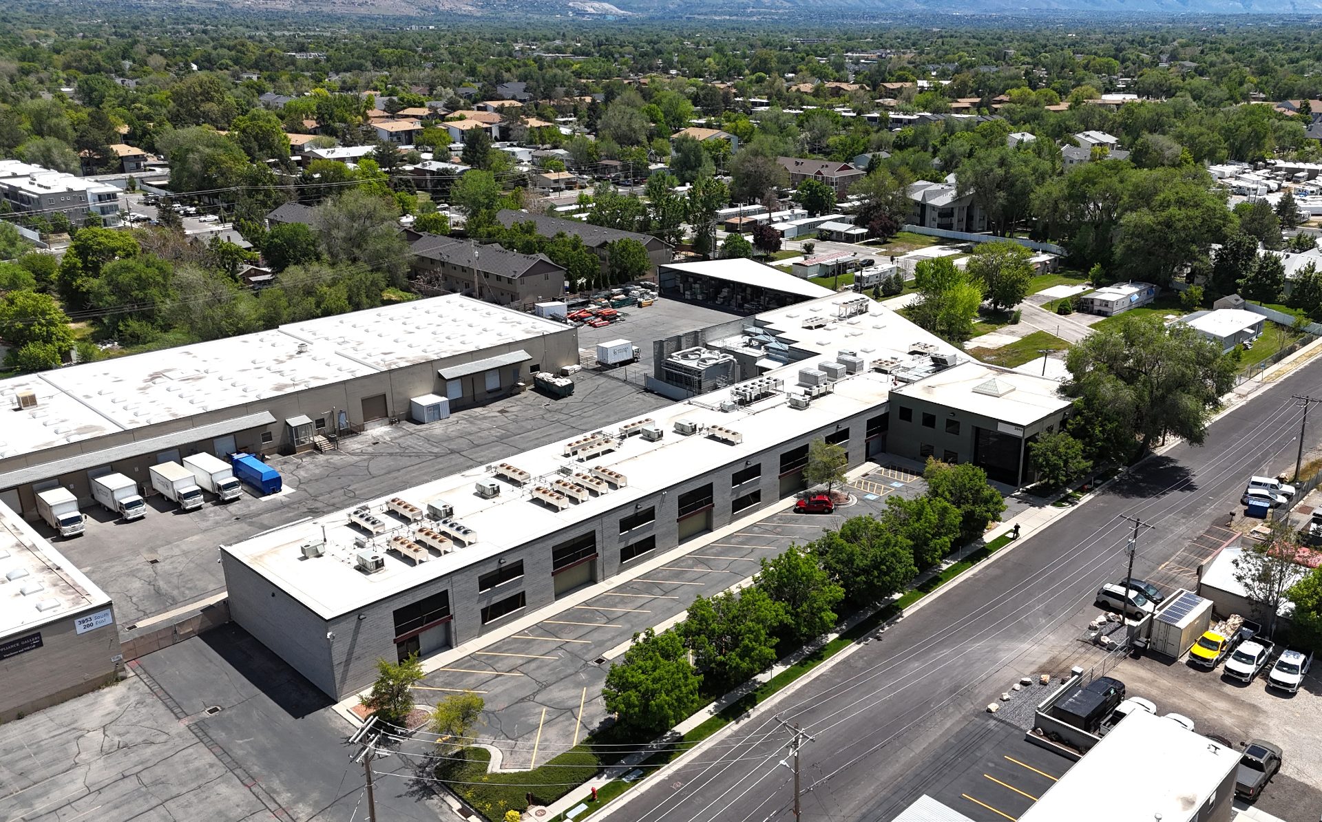 An aerial shot of a long white two-story building. It's in a mostly residential neighborhood, with houses and trees adjacent. Behind it sits a similarly sized buildings that has several trucks parked at bays.