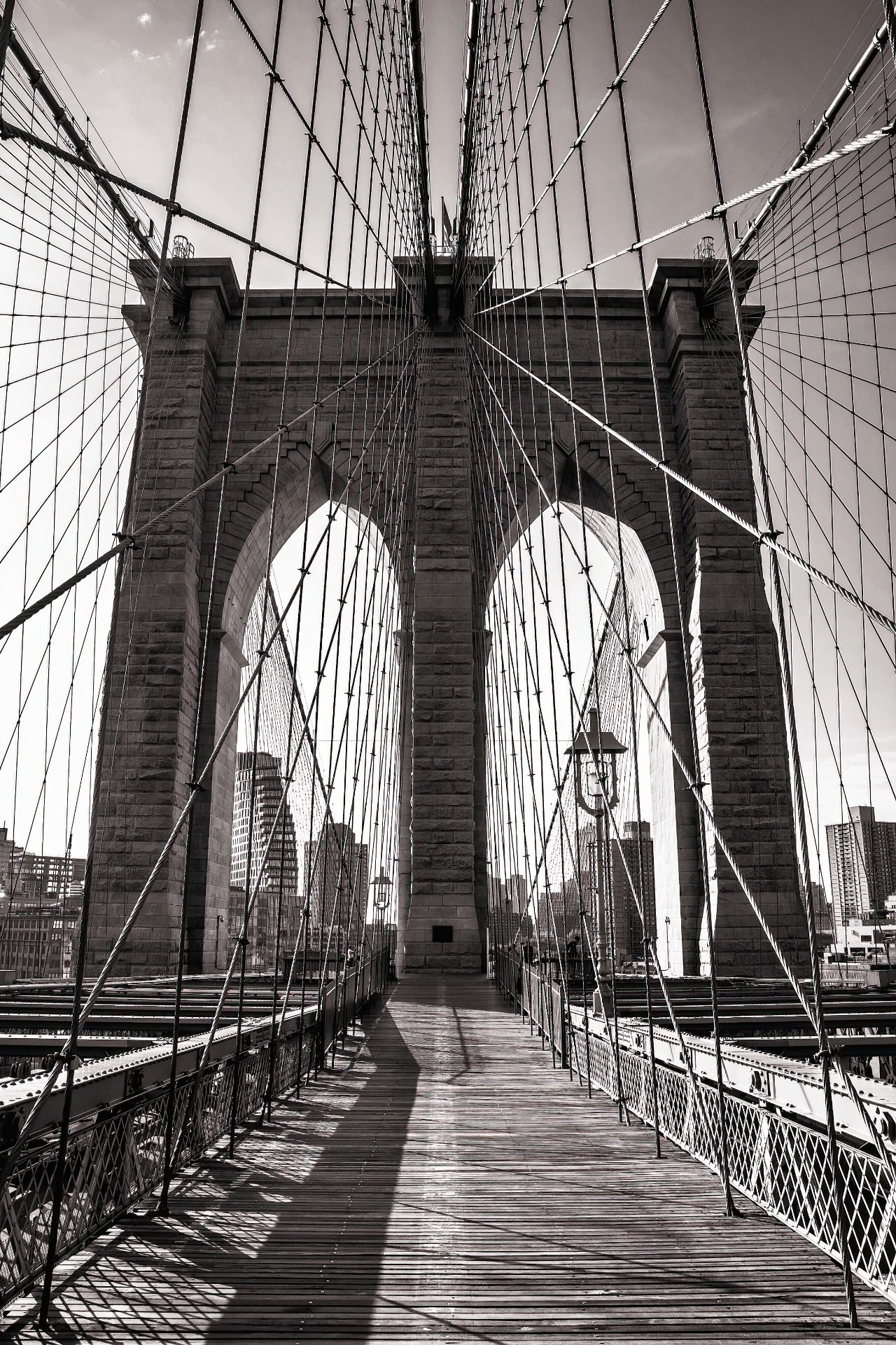 A black-and-white view of a wooden pedestrian walkway on the Brooklyn Bridge that is literally surrounded by suspension wires. Straight ahead is one of the bridge's massive brick suspension pillars. Its twin gothic arches on left and right are pathways that provide access to the rest of the bridge walkway.