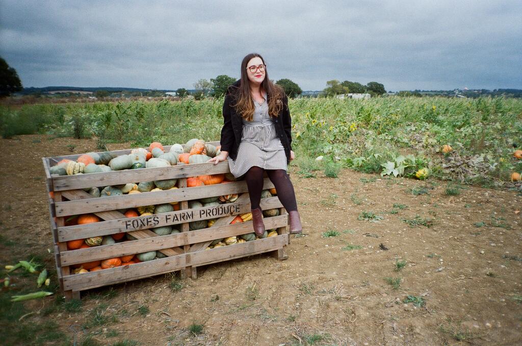 A woman wearing glasses sitting on a crate of pumpkins on a farm