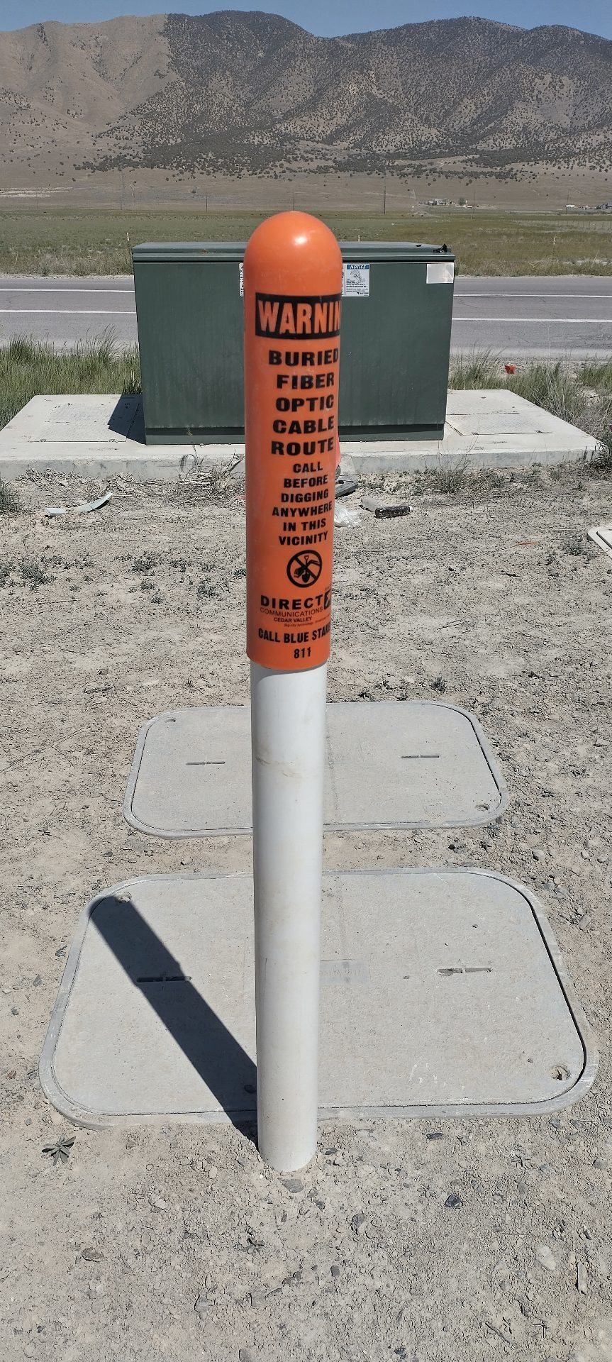 A white pole with an orange cap. The cap reads "WARNING: Buried Fiber Optic Cable Route" and warns the reader not the dig in the area. At its base, one can see an access cover that probably provides access to the fiber.