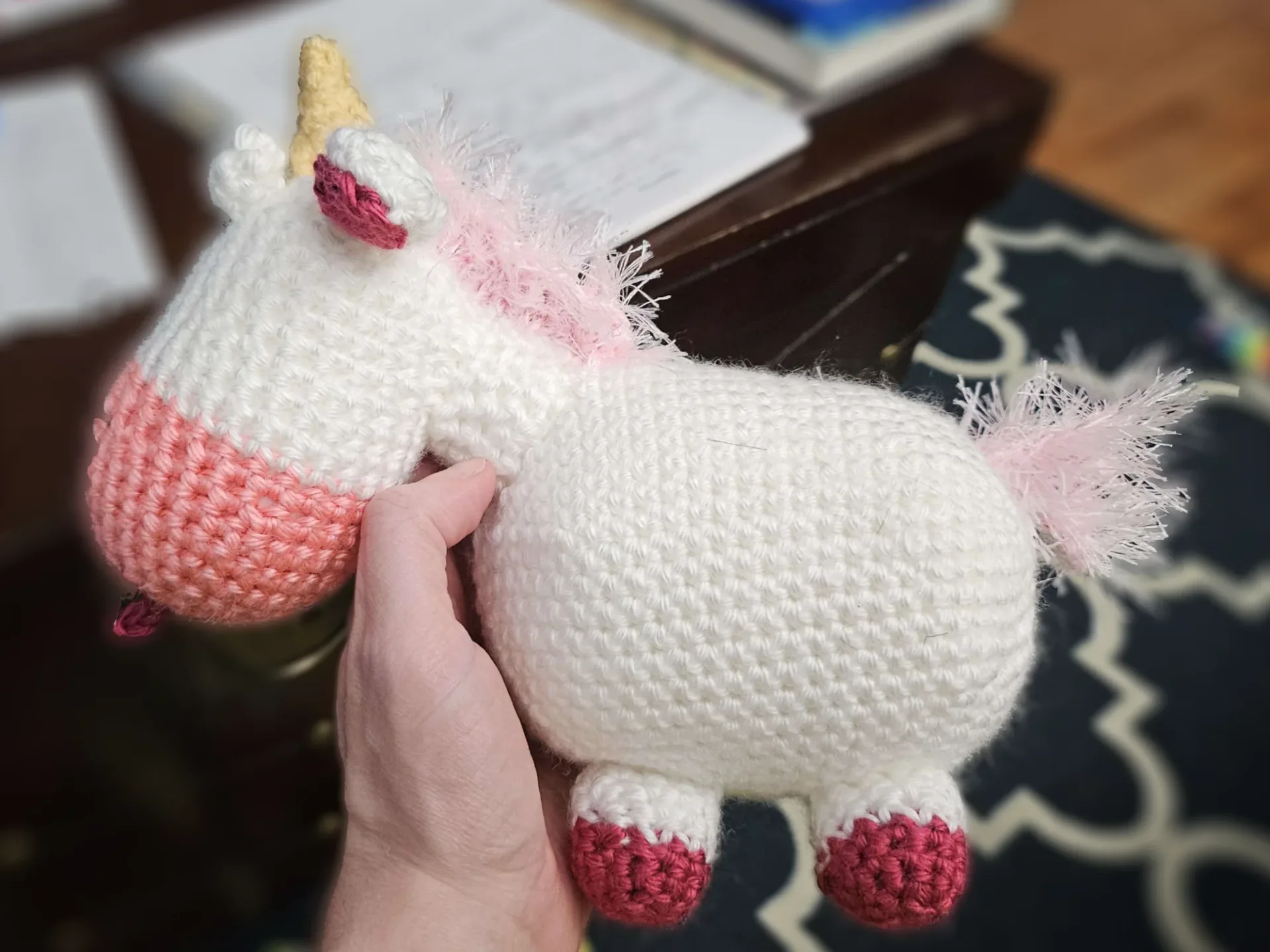 A crocheted unicorn facing the side with bulging eyes and a silly tongue-out smile. It has a pink fluffy mane and tail.