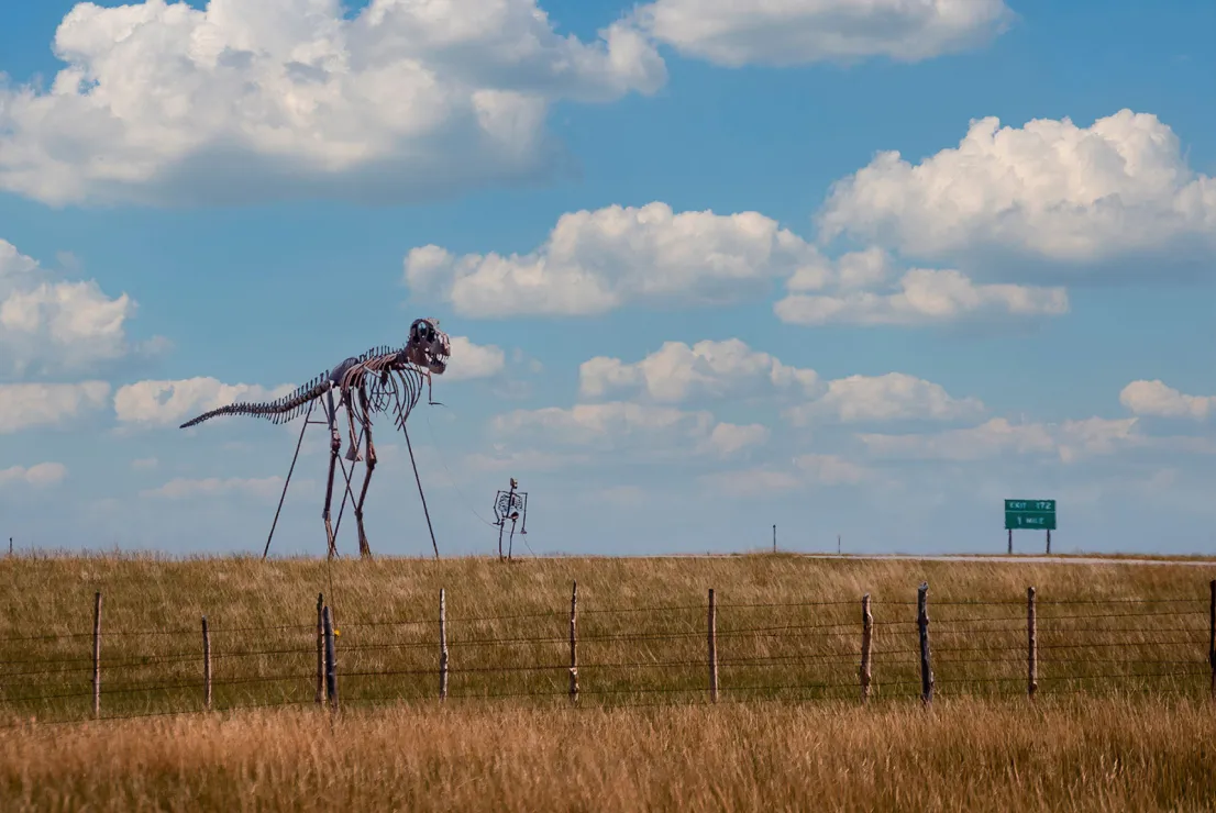 A metal sculpture featuring a skeletal man walking an equally skeletal dinosaur (probably a T-Rex by the size of its tiny little arms) sits beside a barely visible highway. It's pictured from across a grassy field.