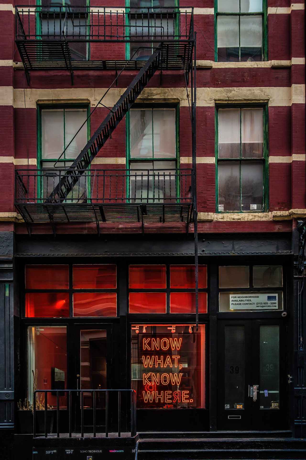 A SoHo building several stories tall has fire escape balconies and stairs running down its red brick exterior. On the bottom level is what appears to be a storefront of some kind, but it's unclear what, if anything, is bought or sold there. In the front window is a orange-red neon sign whose words read "KNOW WHAT, KNOW WHERE." The "r" in "where" is backward and it's a redder colour than the rest of the sign. The interior of the space is lit with an eerie ruby glow. I guess everybody knows that this is nowhere.