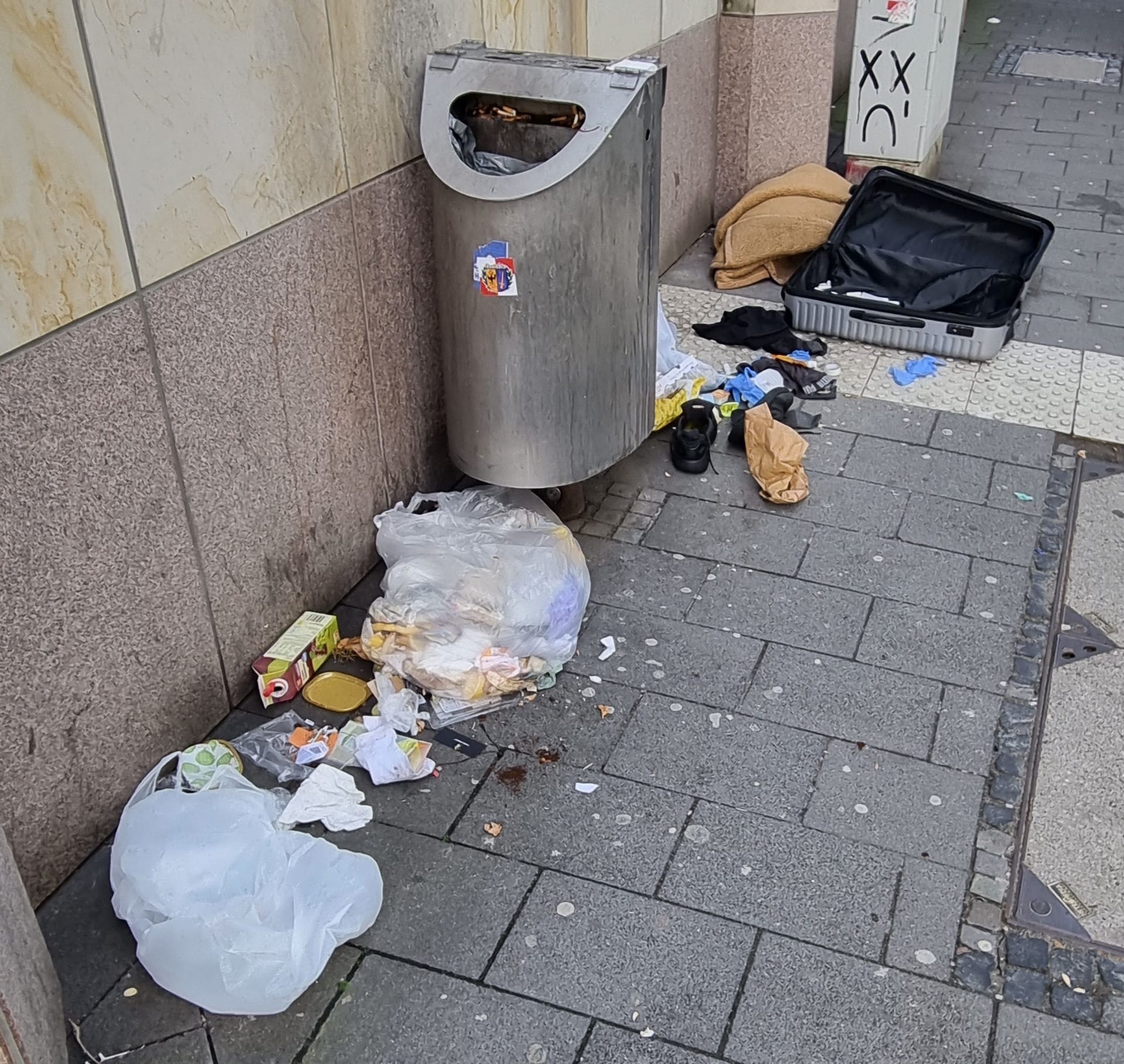 Verschiedener Müll, unter anderem ein Reisekoffer, auf dem Boden vor einem Abfallbehälter.