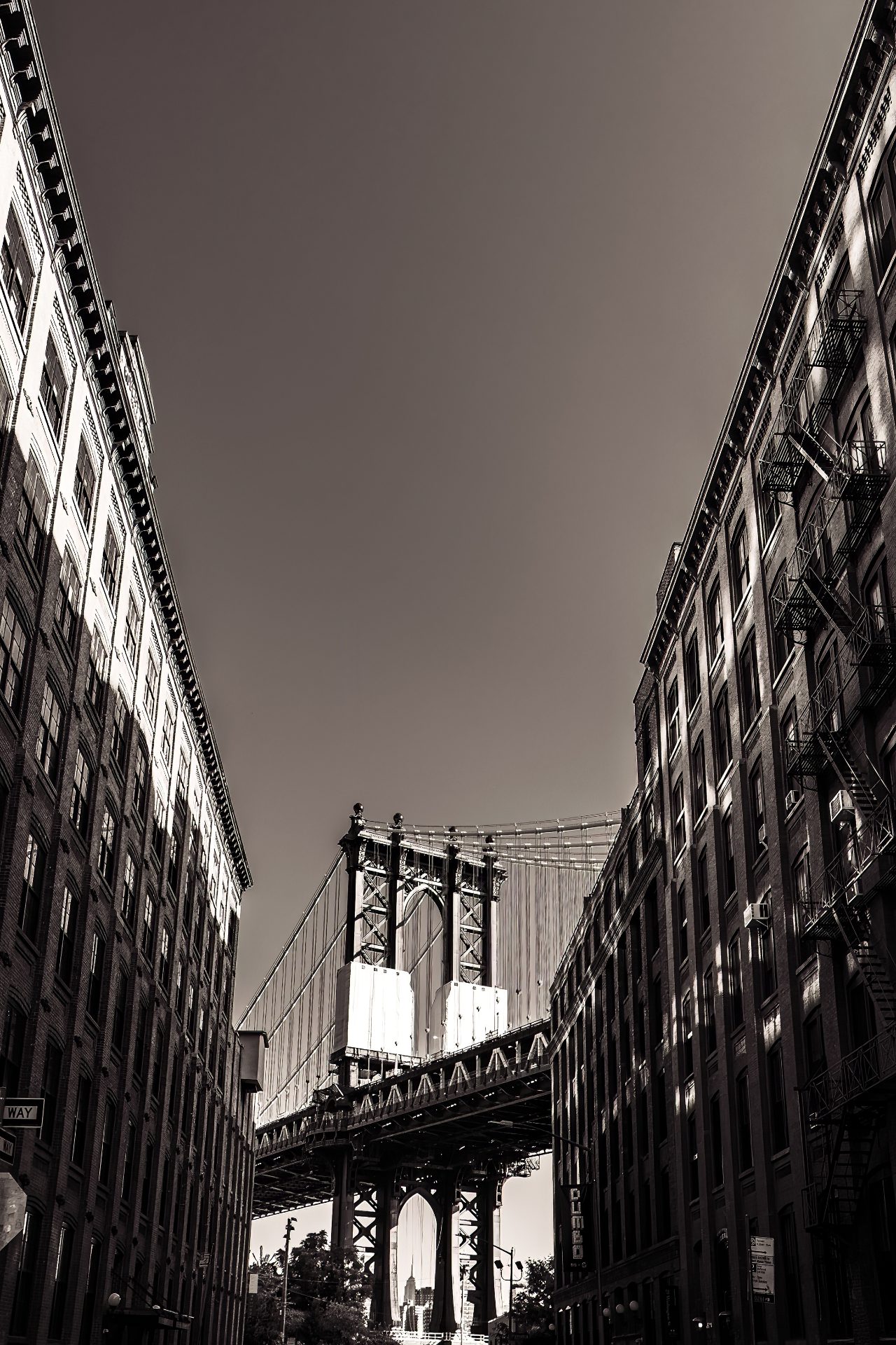 A sepia-toned monochrome depiction of a segment of the Manhattan Bridge as seen from Washington Street in DUMBO, Brooklyn. It's framed between the tallish buildings on either side of the street and the Empire State Building is visible in the distance if you peer through the arch of the bridge support.