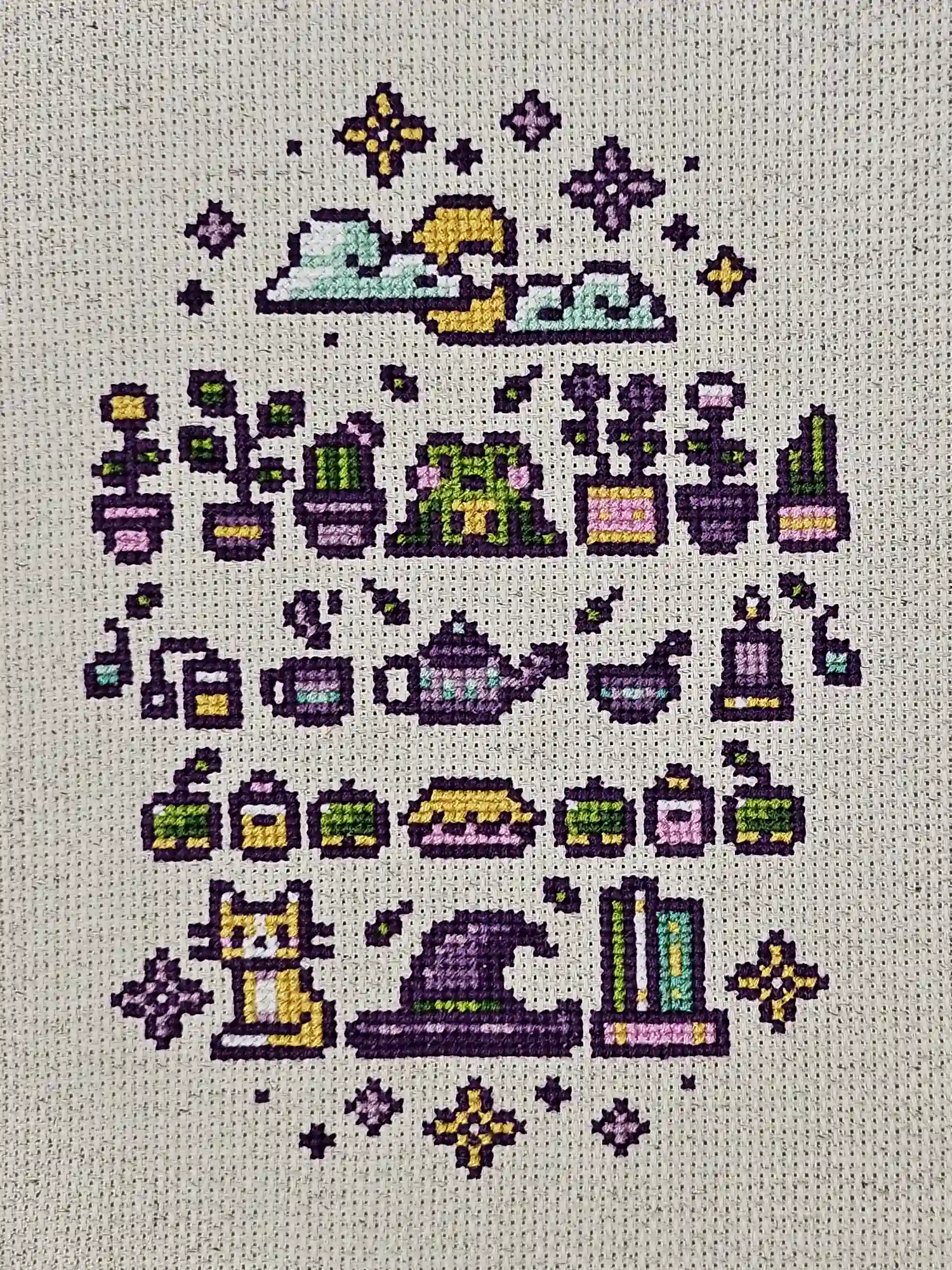 A series of items all fitting a witch's cottage, including plants, a teapot, a frog, and a cat. Cross-stitch.