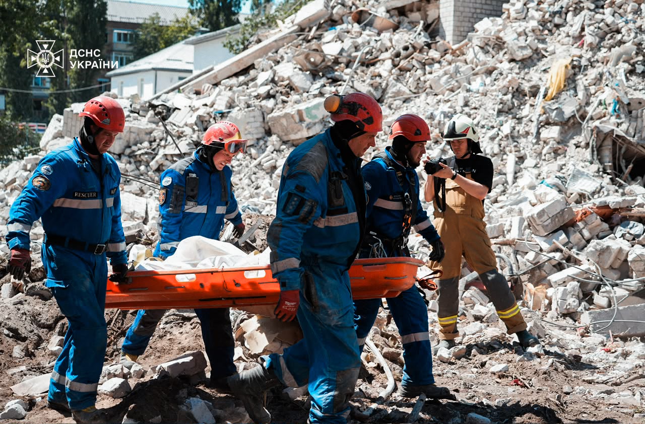 Servicios de emergencia cargando una camilla con un cuerpo envuelto en una bolsa para cadáveres. Caminan sobre los cascotes de un edificio derrumbado por los ataques.