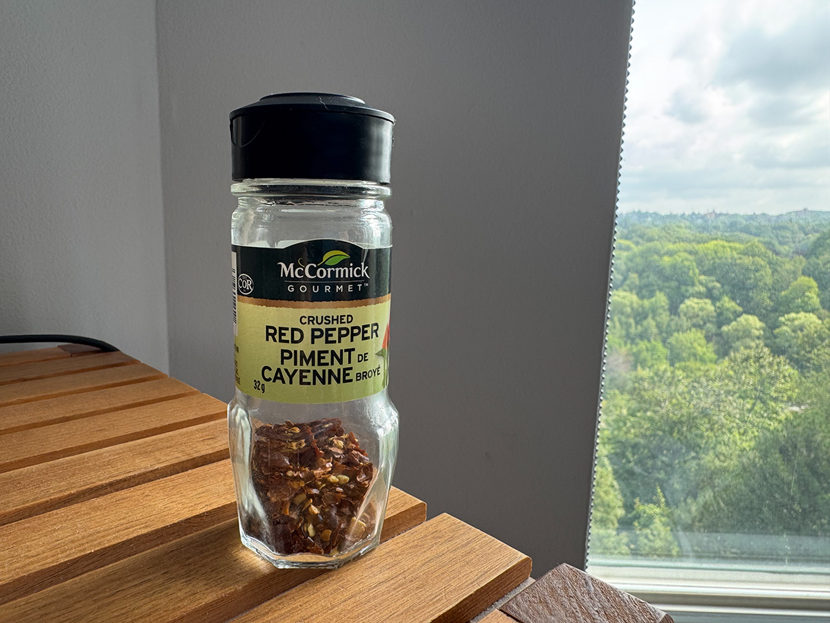 Bottle of McCormick's Crushed Red Pepper, about one third full. Bottle sits on a wooden shelf, window nearby that looks out onto trees.