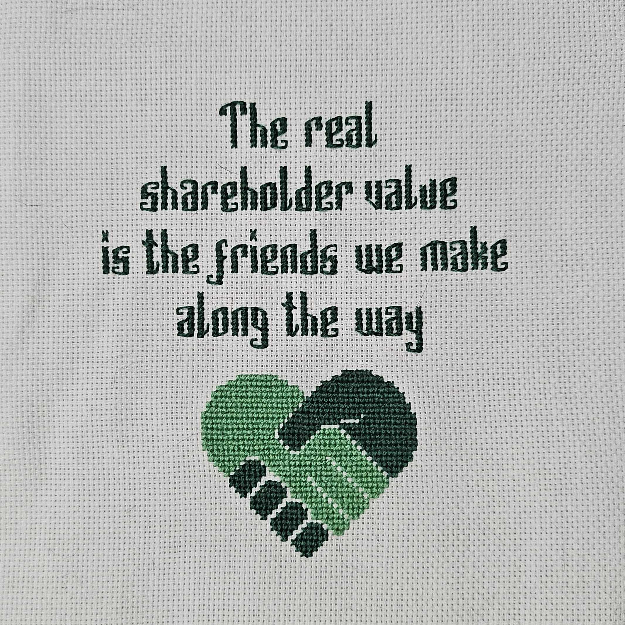 A cross-stitch completed on white aida cloth in teal and green thread. It says, "The real shareholder value is the friends we make along the way" above a pair of hands shaking in a heart shape.