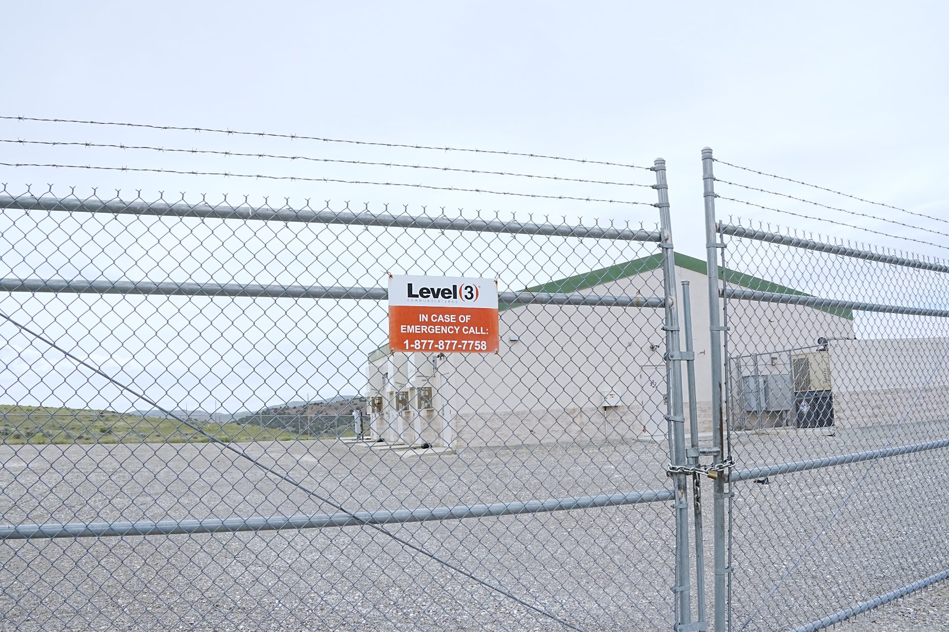A small building behind a chain link fence. On the gate is a sign directing the reader to call Level(3) in an emergency.