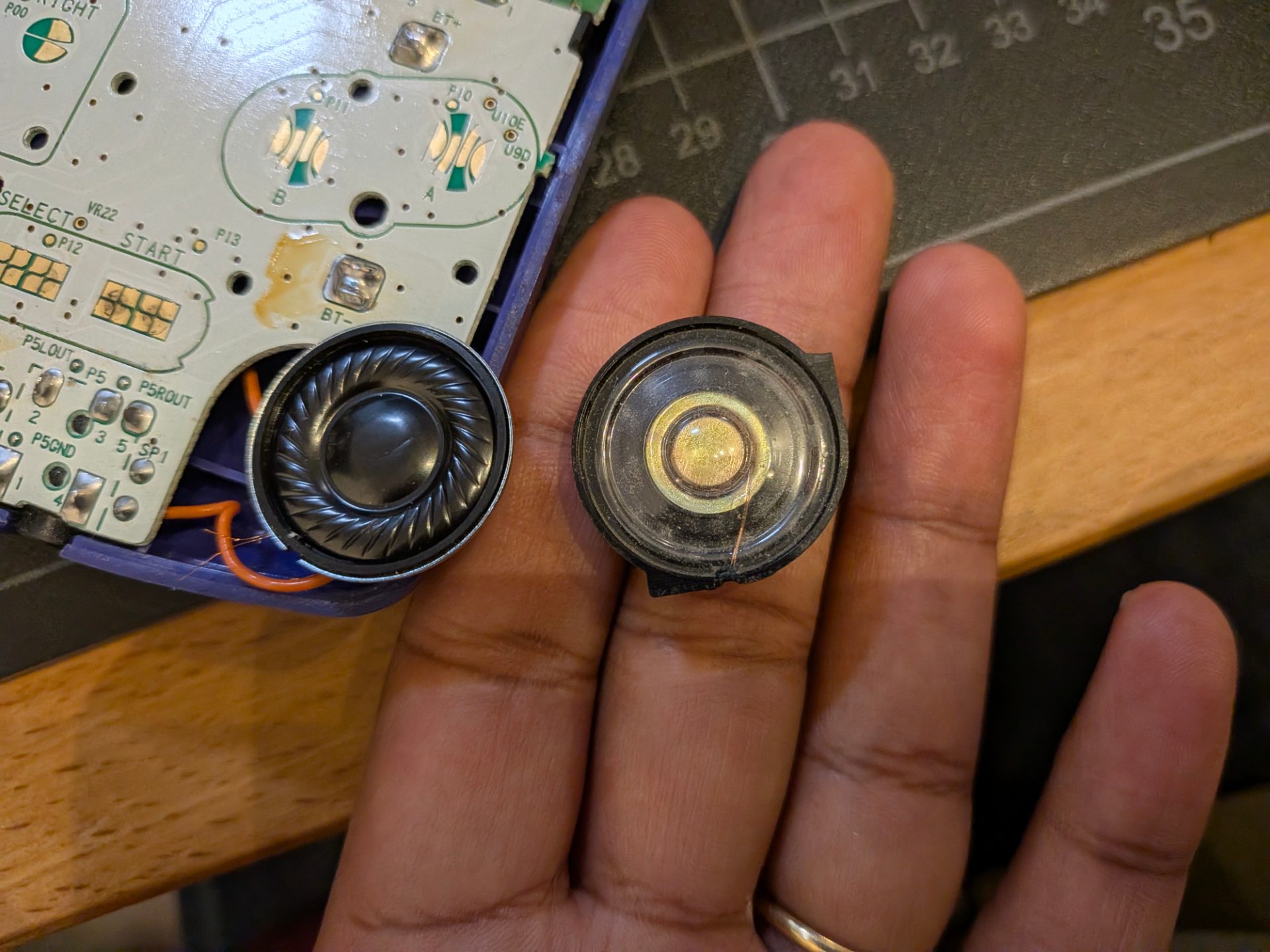 Holding the old speaker next to the new one that has been attached to the circuit board. The old has a thick plastic shell with two wings so it fits into the shell snugly.