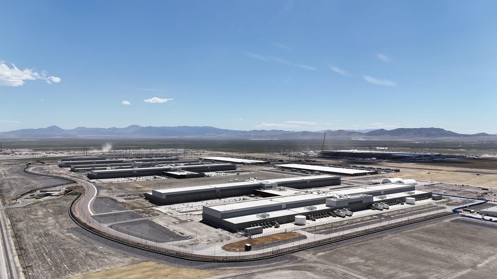 A massive campus stretches out into the distance. In the foreground, two large, long buildings can be seen, surrounded by generation and cooling equipment. The whole campus is ringed by a road.