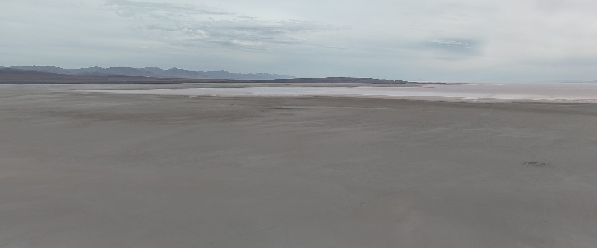 A large expanse of salt flat. It's grey, likely because it was underwater just a few years ago. Further out, a bit of water can be seen. It's pinkish - not an illusion, but a result of minerals and a specific type of high-alkalinity algae.