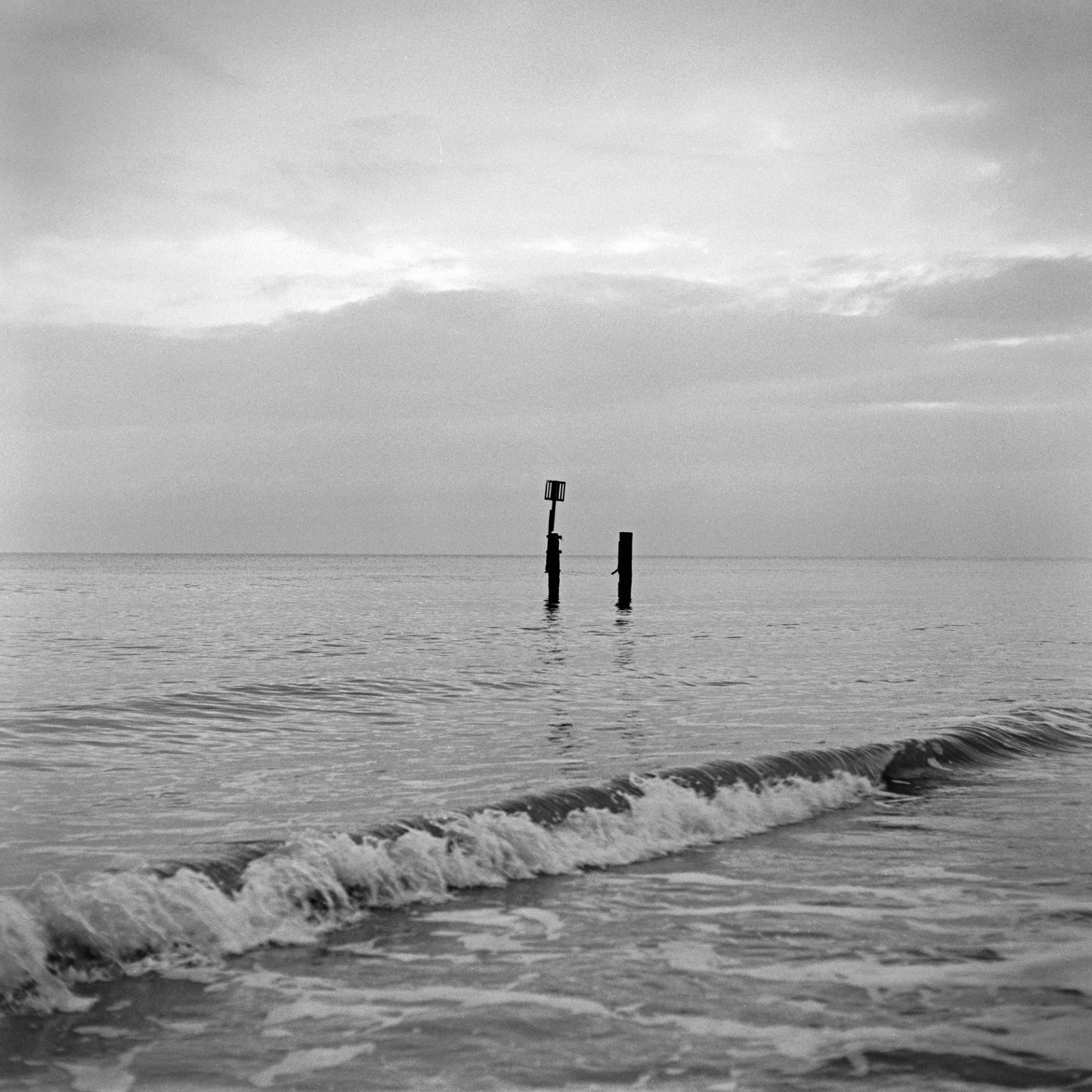 A black and white square photograph captures a calm sea under an overcast sky, with gentle waves rolling onto the shore in the foreground. In the middle ground, two dark wooden groyne posts emerge from the calm water, one of which is topped with a square navigational marker. The horizon line sits low in the frame, separating the still ocean from a dense, textured layer of grey clouds.