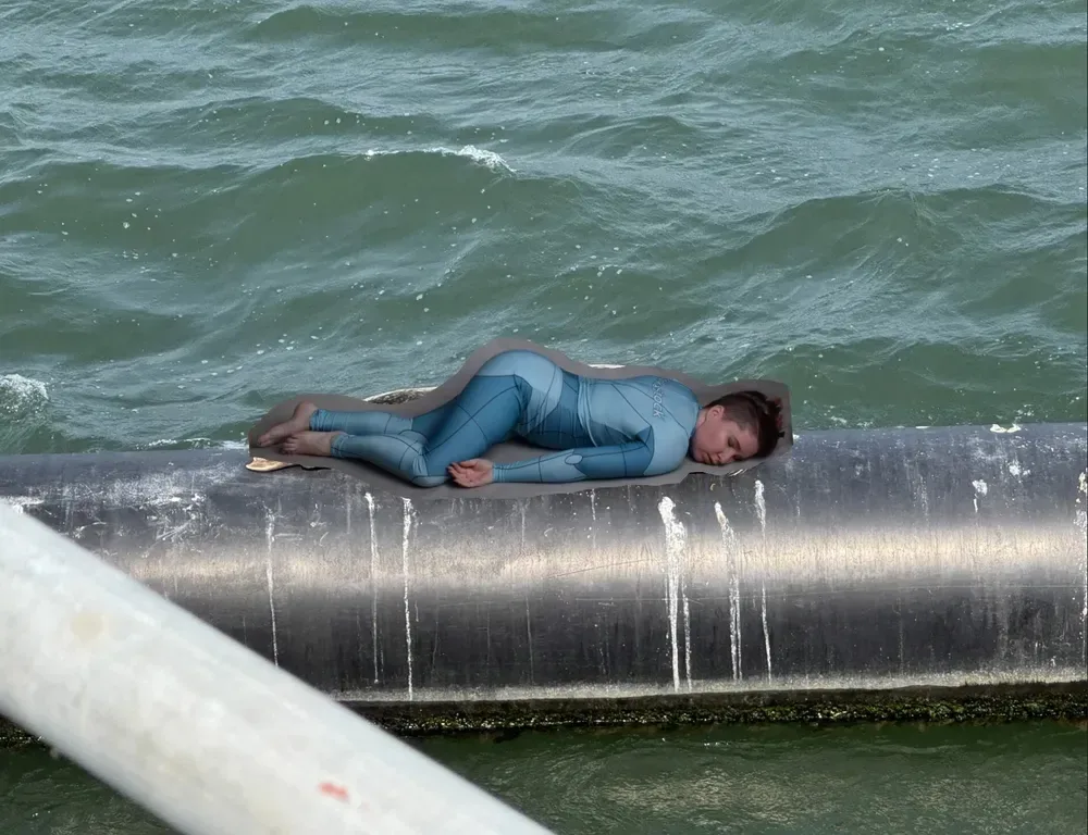 a light skinned, dark haired woman is laying face down. She is poorly photoshopped onto a tube/pipe floating in the ocean water