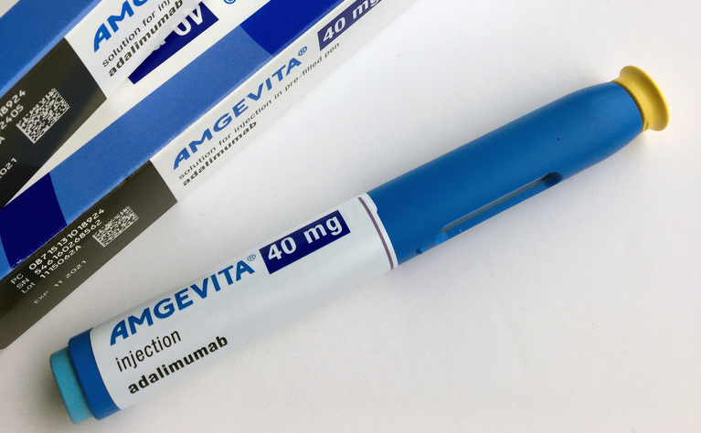 In the top left, the image shows two boxes of Amgevita 40mg boxes in a blue & white colour scheme.
Going diagonally from bottom left to top right is a self-injector pen of 40m of Amgevita (adalimumab). The pen is royal blue in colour with a pale blue button at the bottom, and a yellow safety cap at the injection point.
The slit for viewing the liquid is just visible near the yellow-capped end.