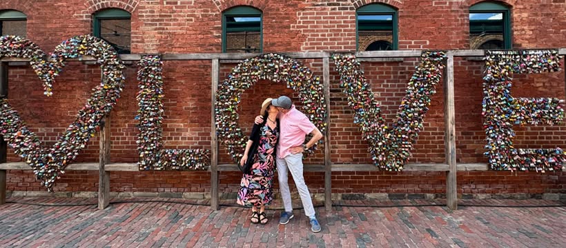 Two people stand in front of the large LOVE sculpture in Toronto's Distillery District, kissing. The letters are covered with colourful padlocks, with a red brick building in the background.