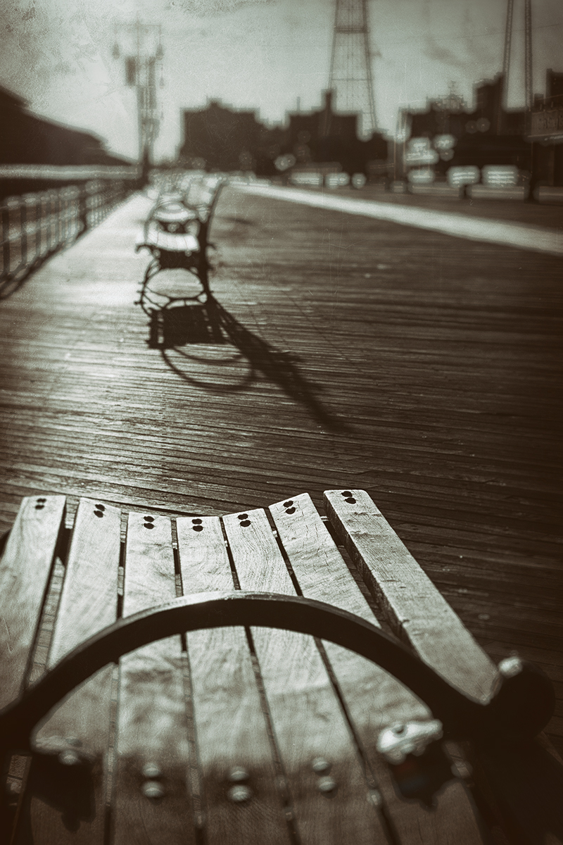 A sepia-toned old-timey rendering of the boardwalk at Coney Island in Brooklyn, New York. By the shadows, you can see that the sun is going down. The bench in the foreground closest to the camera is in focus, but you can see a row of other benches marching off into the distance, all empty. There's absolutely no-one visible on the boardwalk at all. Not surprising, as it's March.