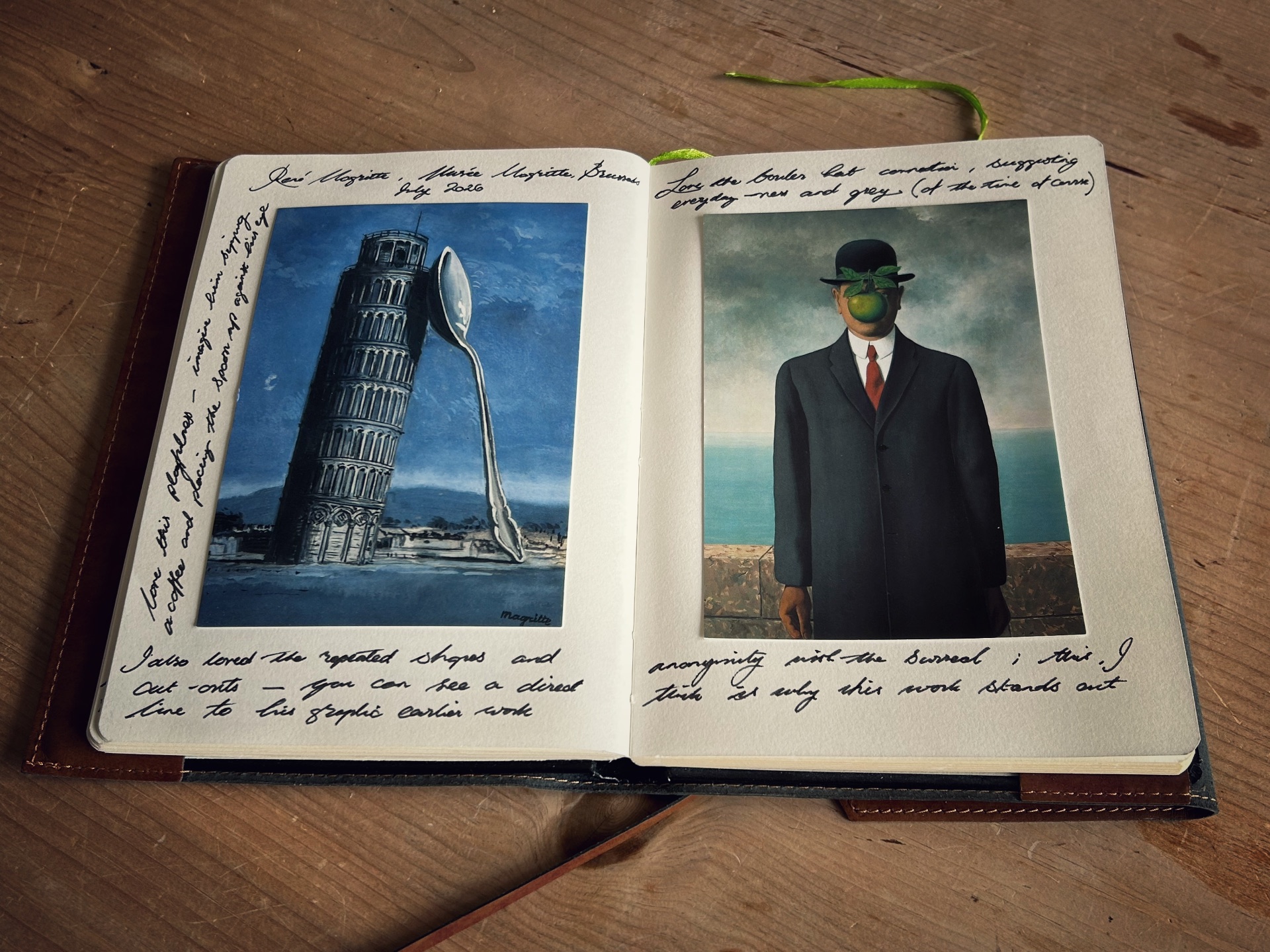 An open sketchbook with leather casing rests on a wooden surface, displaying postcards and handwritten notes about Belgian surrealist painter René Magritte.The left page features a postcard of Magritte’s painting The Labors of Alexander, depicting the Leaning Tower of Pisa propped up by a giant silver spoon under a blue sky. Handwritten notes curve around the image and line the bottom of the page.The right page holds a postcard of The Son of Man, showing a man in a dark overcoat and bowler hat with his face obscured by a floating green apple against a cloudy sky and sea backdrop. Additional cursive handwriting fills the space above and below the postcard.