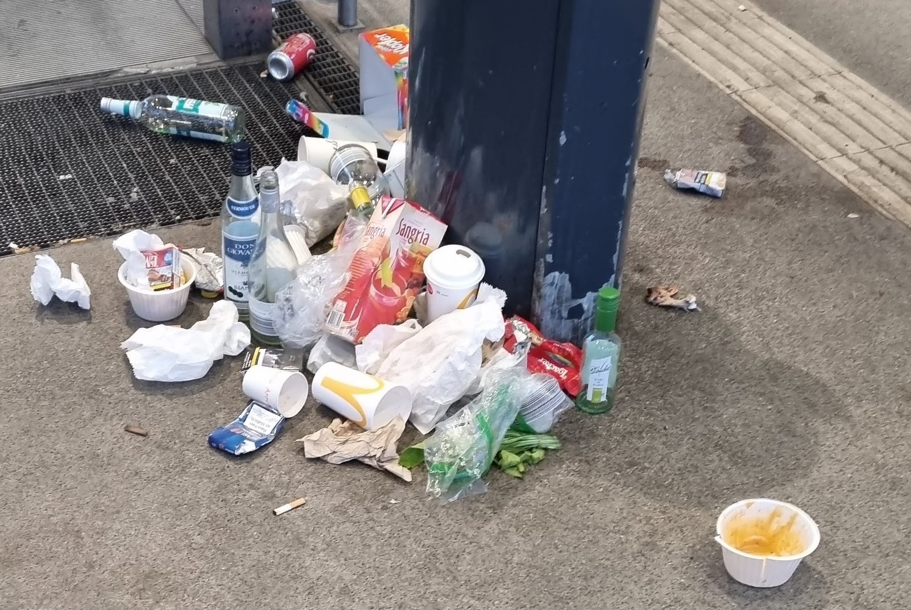 Verschiedene Abfälle, unter anderem Verpackungen von Schnellrestaurants, auf dem Boden vor einem Mülleimer an einer Stadtbahnhaltestelle.