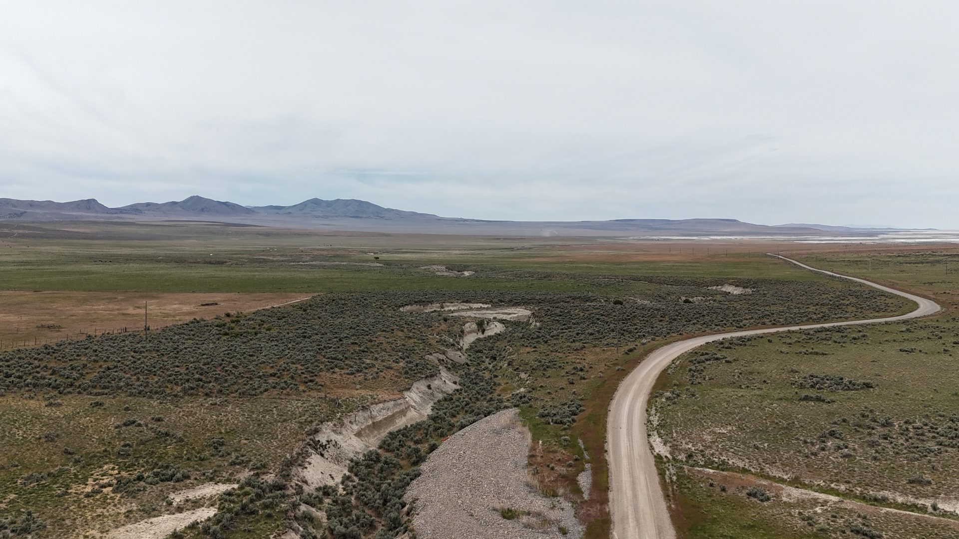 A wide aerial shot. We can see a fairly flat valley, some of it appears to be irrigated and green, other parts seem brown. A road snakes through the picture roughly 2/3 of the way to the right. A wash gully threads it way through the center. In the background on the left are mountains, and on the right, it's salt flats.