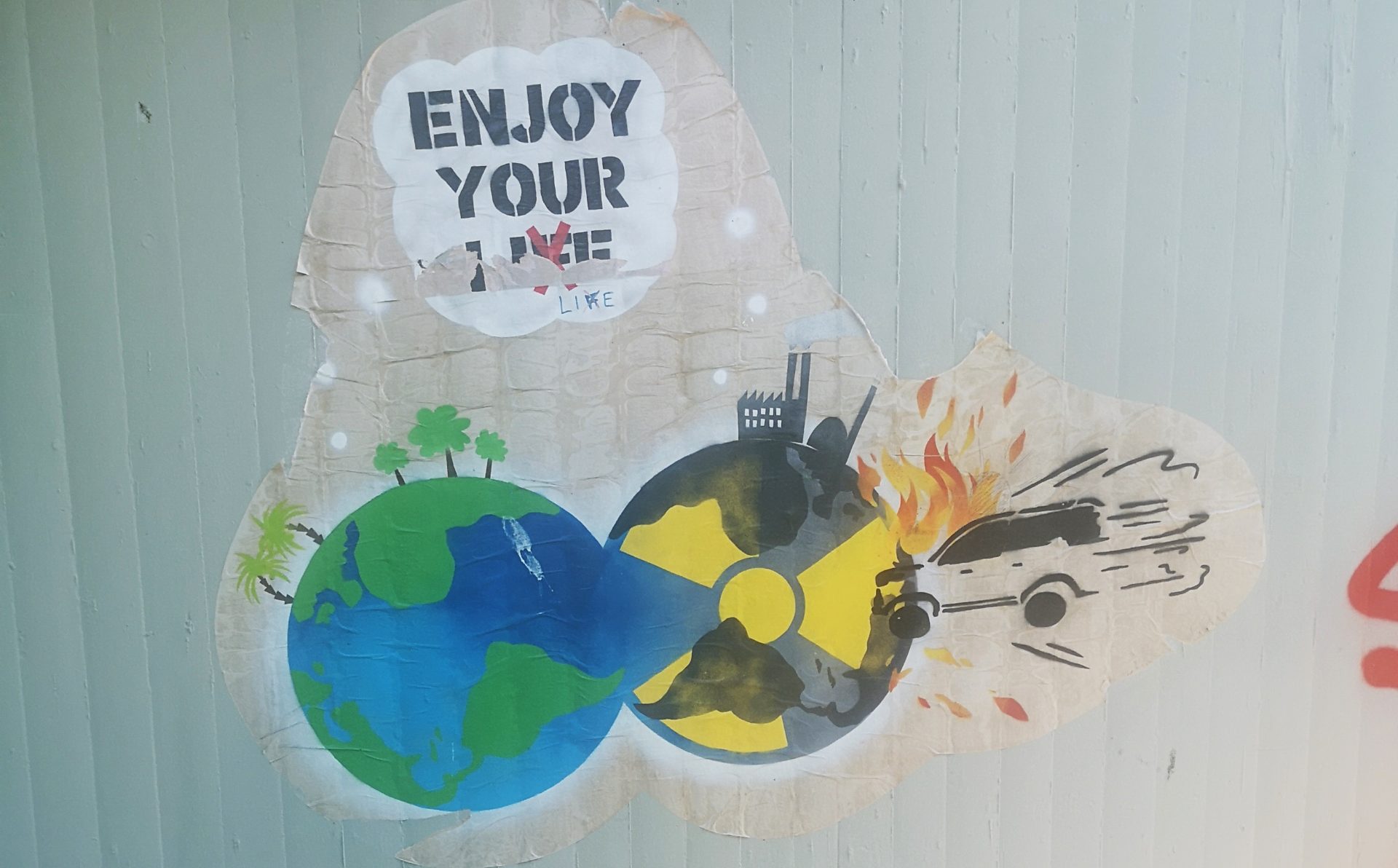 Paste-Up auf einer Betonwand. Eine Sprechblase mit den Worten enjoy your life. Das F in life ist mit einem roten Kreuz durchgestrichen. Darunter zwei nebeneinanderliegende Erdkugeln. Die linke mit blau, für das Wasser und grün, für die Landmasse. Auf ihr zwei Palmen und drei Bäume. Die rechte mit dem Warnzeichen für Radioaktivität. Ein Fabrikgebäude und ein Kernkraftwerk stehen darauf. Ein Auto das in die Kugel krachte und aus dessen Motor Flammen schlagen.