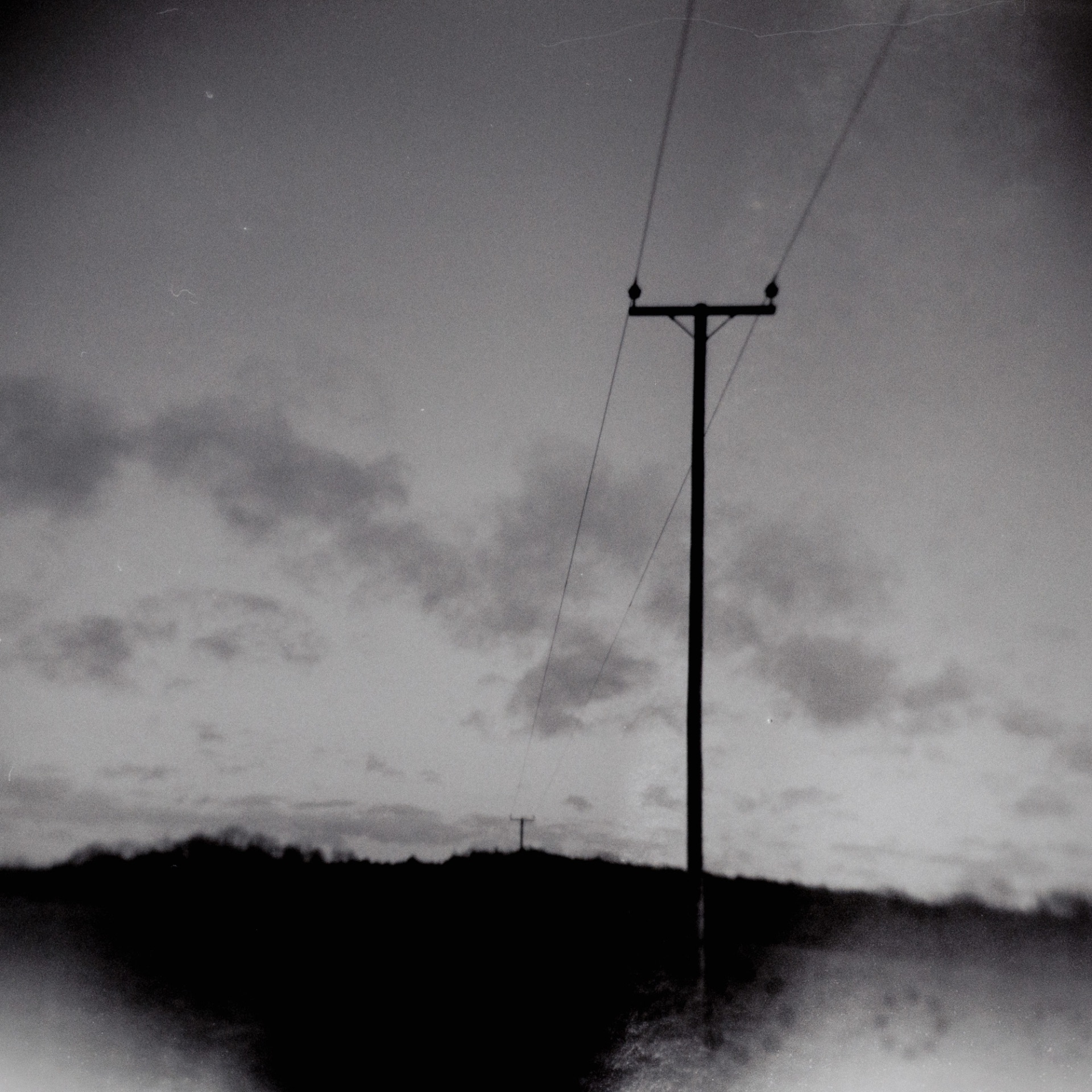 A grainy, black and white square Holga photograph shows a tall wooden telegraph pole standing prominently on the right side of the frame. Cable lines stretch from its T-shaped crossarm toward the upper right corner and down toward a distant hill. The horizon is a dark, blurry silhouette of a hill with a second, tiny telegraph pole visible on the ridge line. The sky is filled with soft, scattered clouds, and the bottom corners of the image feature heavy vignetting and soft-focus blurring.