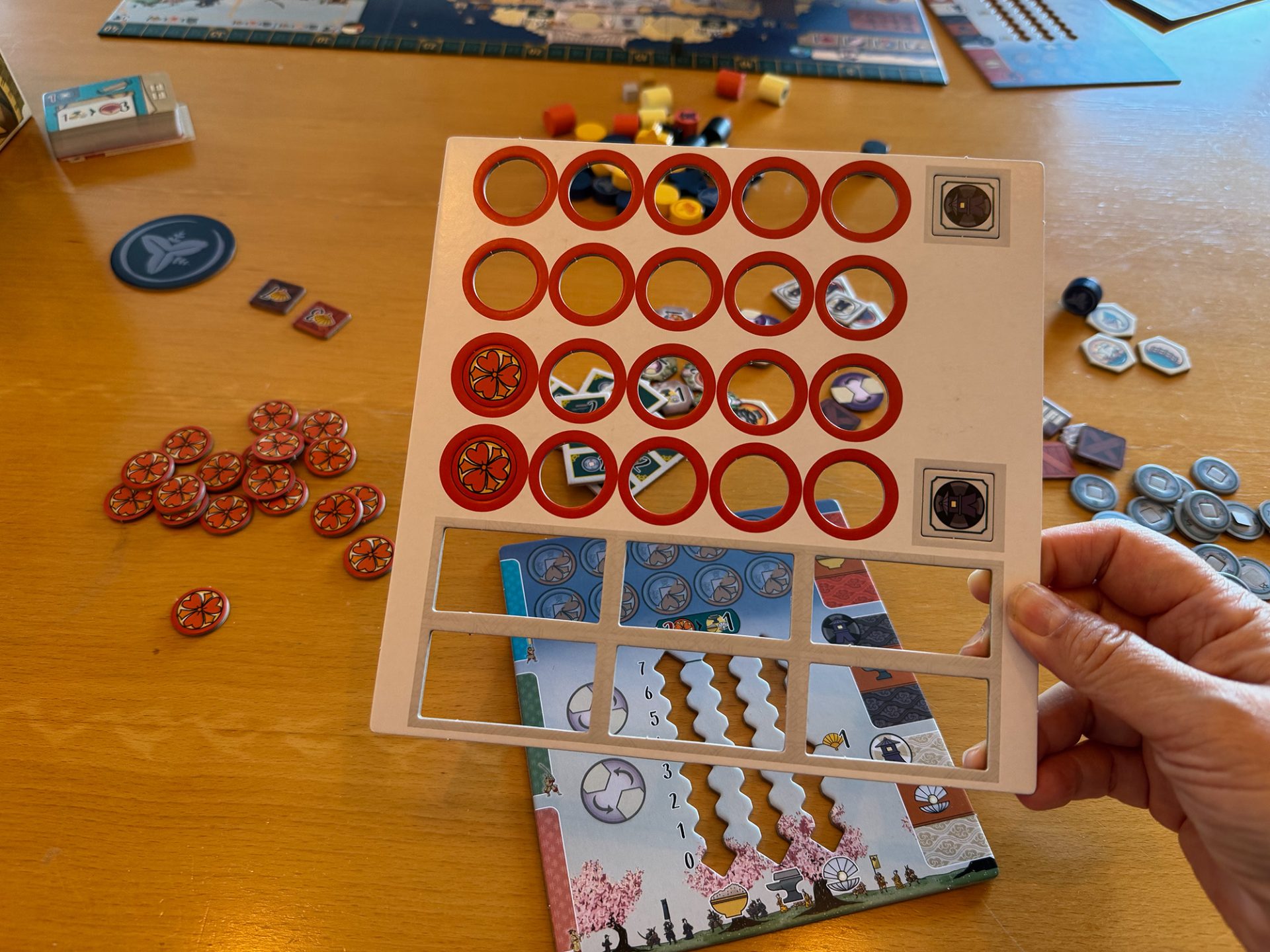 A hand holds an empty cardboard punchboard frame from a board game called White Castle, above a table scattered with orange, blue, and other game tokens and player boards.