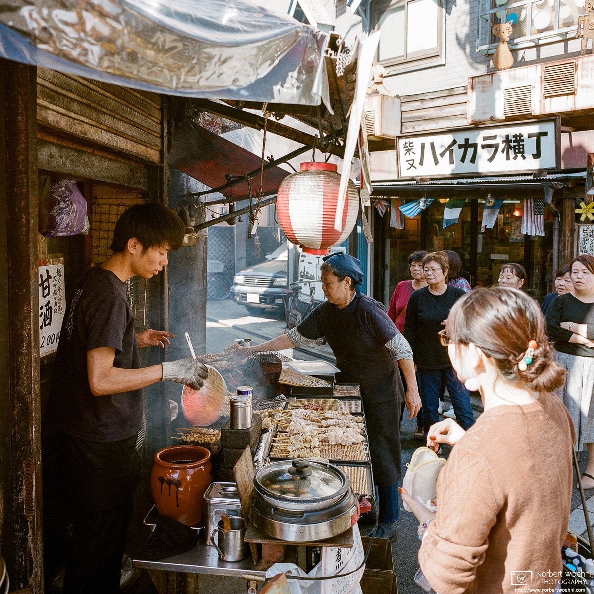 People are watching food being prepared at a Yakitori (skewered charcoal-grilled chicken) stand in Shibamata, Tokyo.