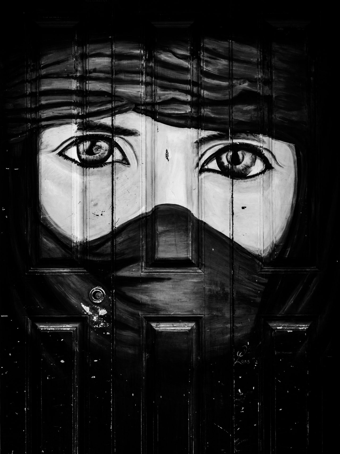 A high-contrast, monochrome artwork painted directly onto the panels of a dark wooden door. The center features a close-up portrait of two expressive eyes and eyebrows, with the rest of the face and head wrapped in a veil. A keyhole lock is visible on the lower left side of the door.