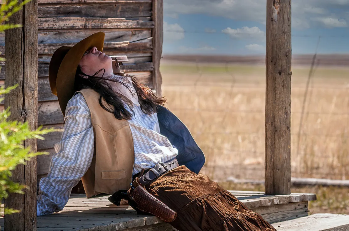 A woman dressed in old-timey Western gear, including a gun strapped to her right hip, sits and leans back on the steps of a covered porch of a wooden house. Her hat is tipped FAR forward, covering her eyes as she relaxes in the shade.