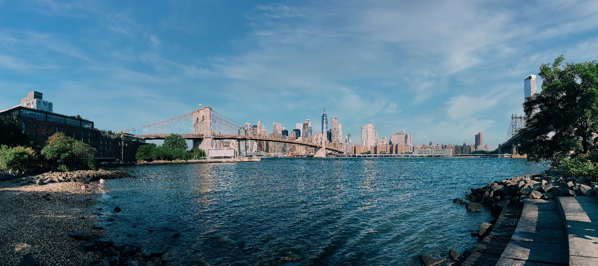 A wide panoramic view of Lower Manhattan from Brooklyn Bridge Park shows the Brooklyn Bridge on the left, the Manhattan Bridge on the right, and across the East River, the tall buildings of the Financial District, One World Trade Centre, and the Two Bridges area of Manhattan.