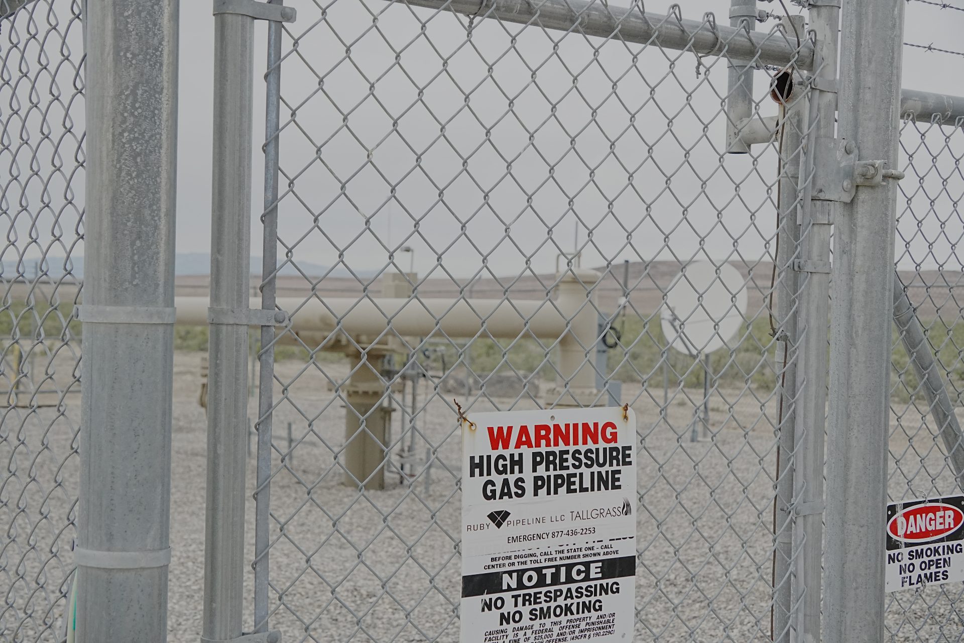 A natural gas pipeline station, surrounded by fending. One can see a large pipeline coming up through the ground, making a horizontal curve, then going back underground. Other equipment inside the fence includes onsite power generation  and telecommunications equipment.