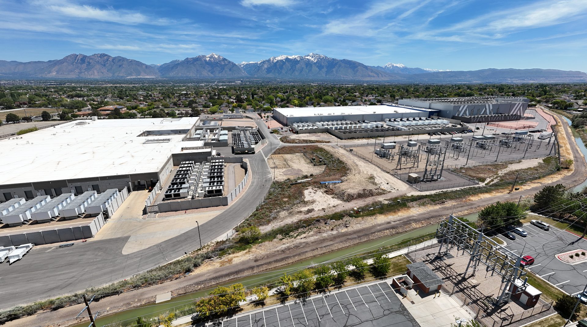 A wide shot of a datacenter campus; three large buildings can be seen. The one on the left has a flat roof and is surrounded by cooling and generation equipment. The one near the center looks similar. The one to the right is twice as tall as the others, and has its equipment mounted on the roof. Power lines, transformers, etc can be seen on the right and in the foreground. In the background, the Wasatch Mountains still have some snow.