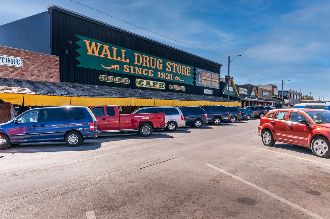 A row of stores on a street is dominated by a large building: this, according to its sign, is the Wall Drug Store (since 1931) offering a cafe, an art gallery, a pharmacy, a museum, and a chapel (among other things). Rows of cars are parked diagonally in front of and across the street from the buiding.