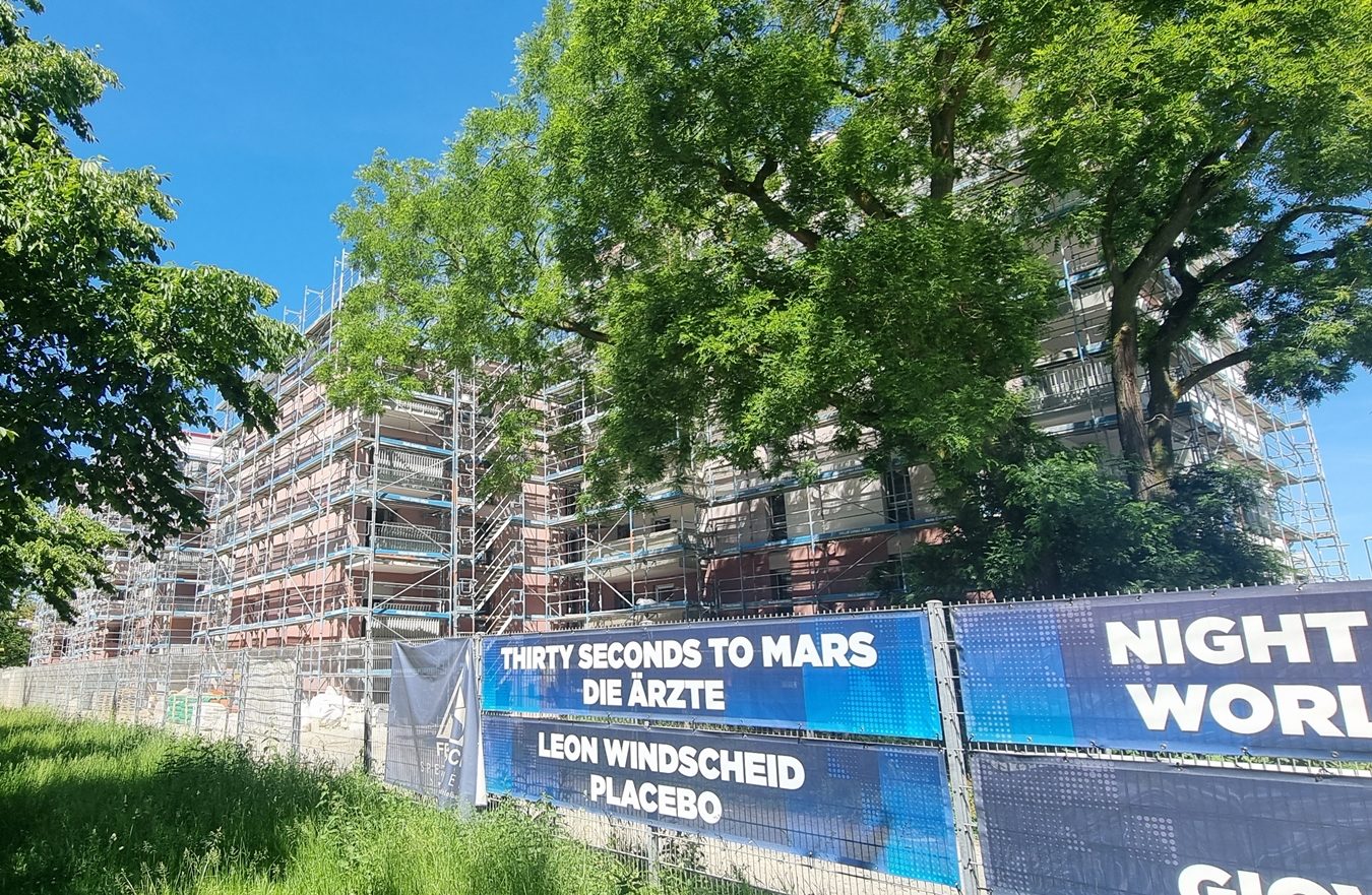 Eingerüsteter Neubau eines Mehrparteienhaus. Verdeckt durch zwei Bäume. Im Vordergrund ein Zaun mit Planen die auf Veranstaltungen bewerben.